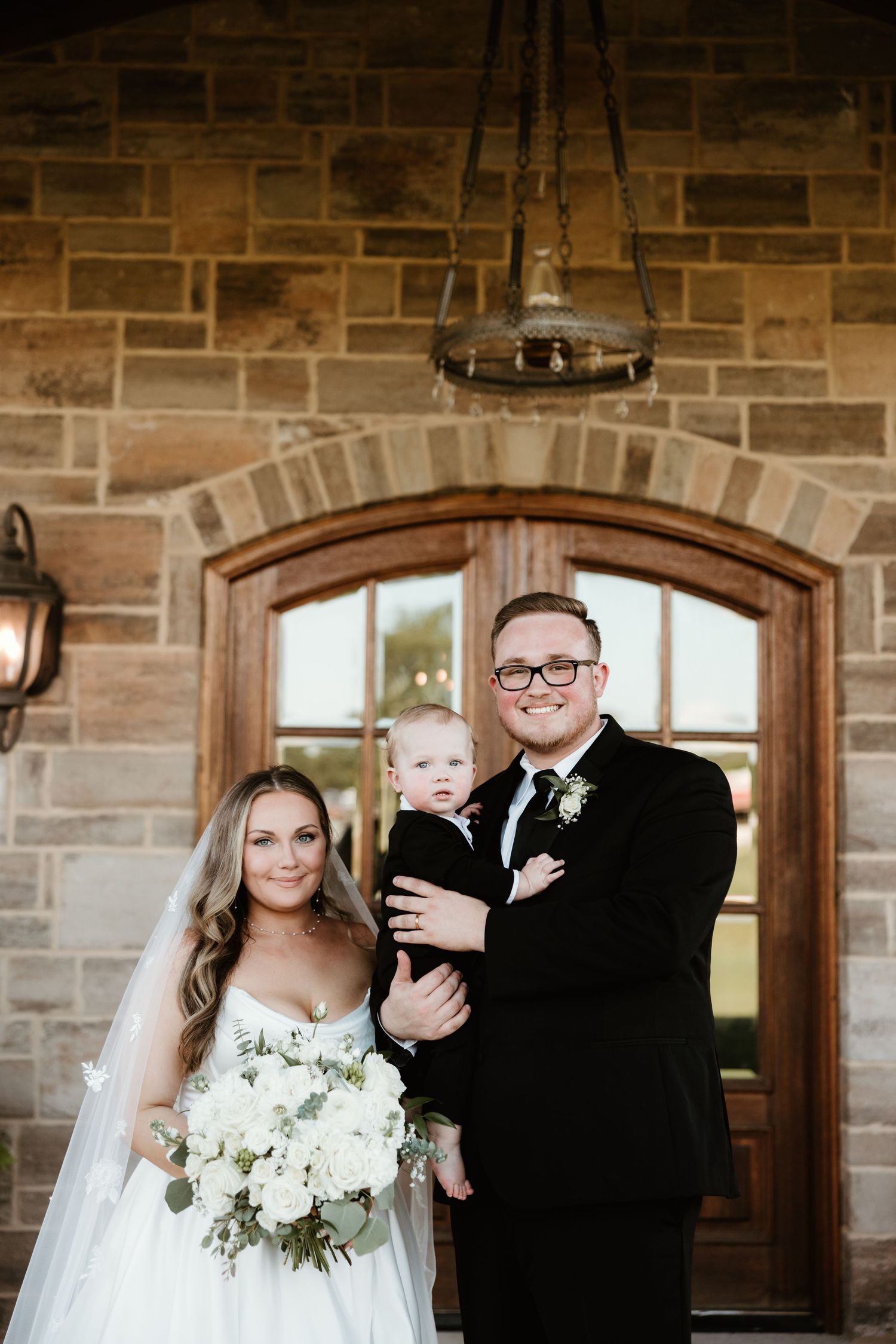 Elegant wedding portrait in front of stone building with arched doorway and rustic chandelier.