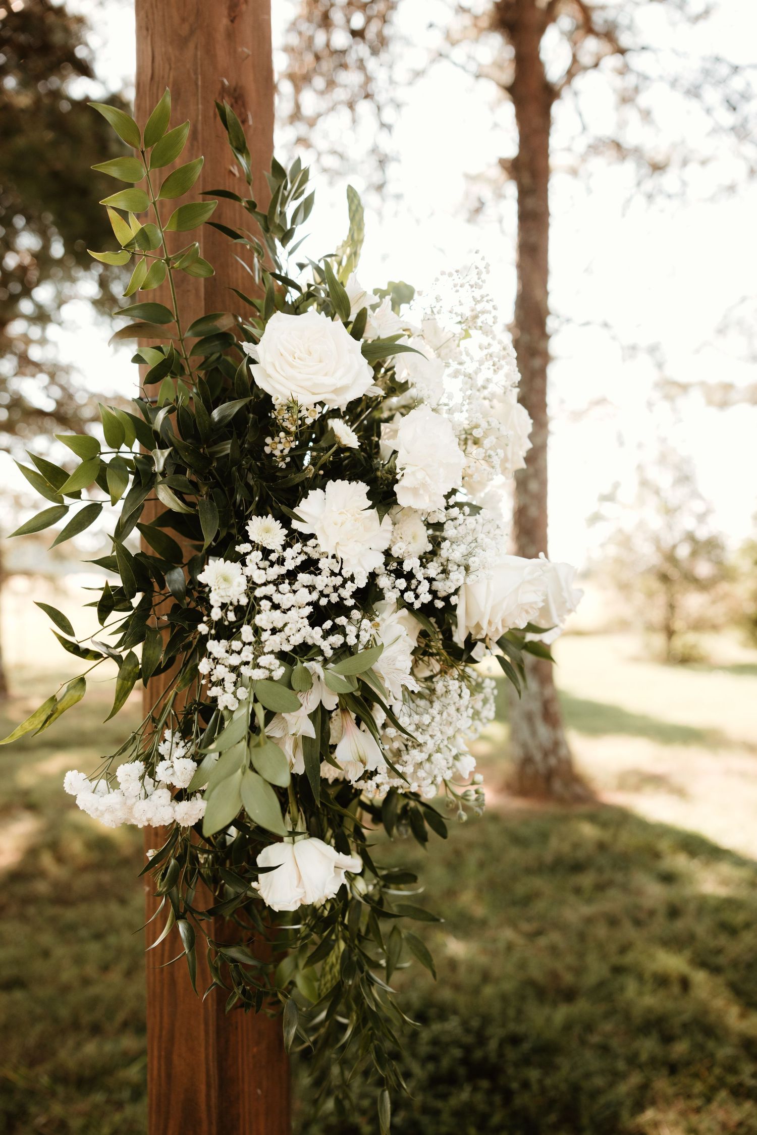 Elegant white floral arrangement with greenery decorates a wooden post in an outdoor setting during golden hour.