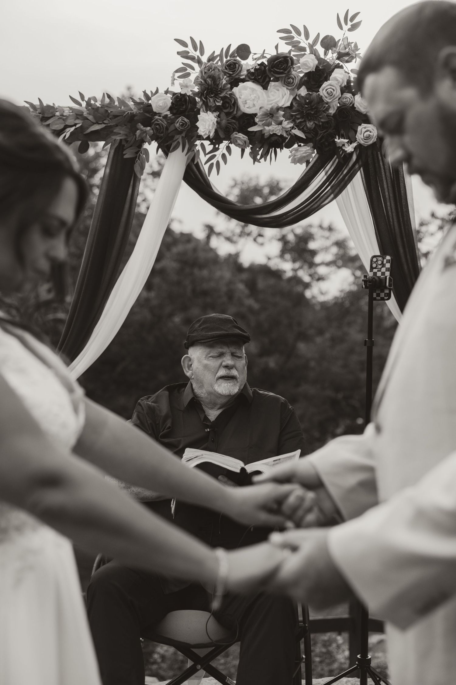 A black and white wedding ceremony photograph showing a floral arch with an officiant conducting the service outdoors.