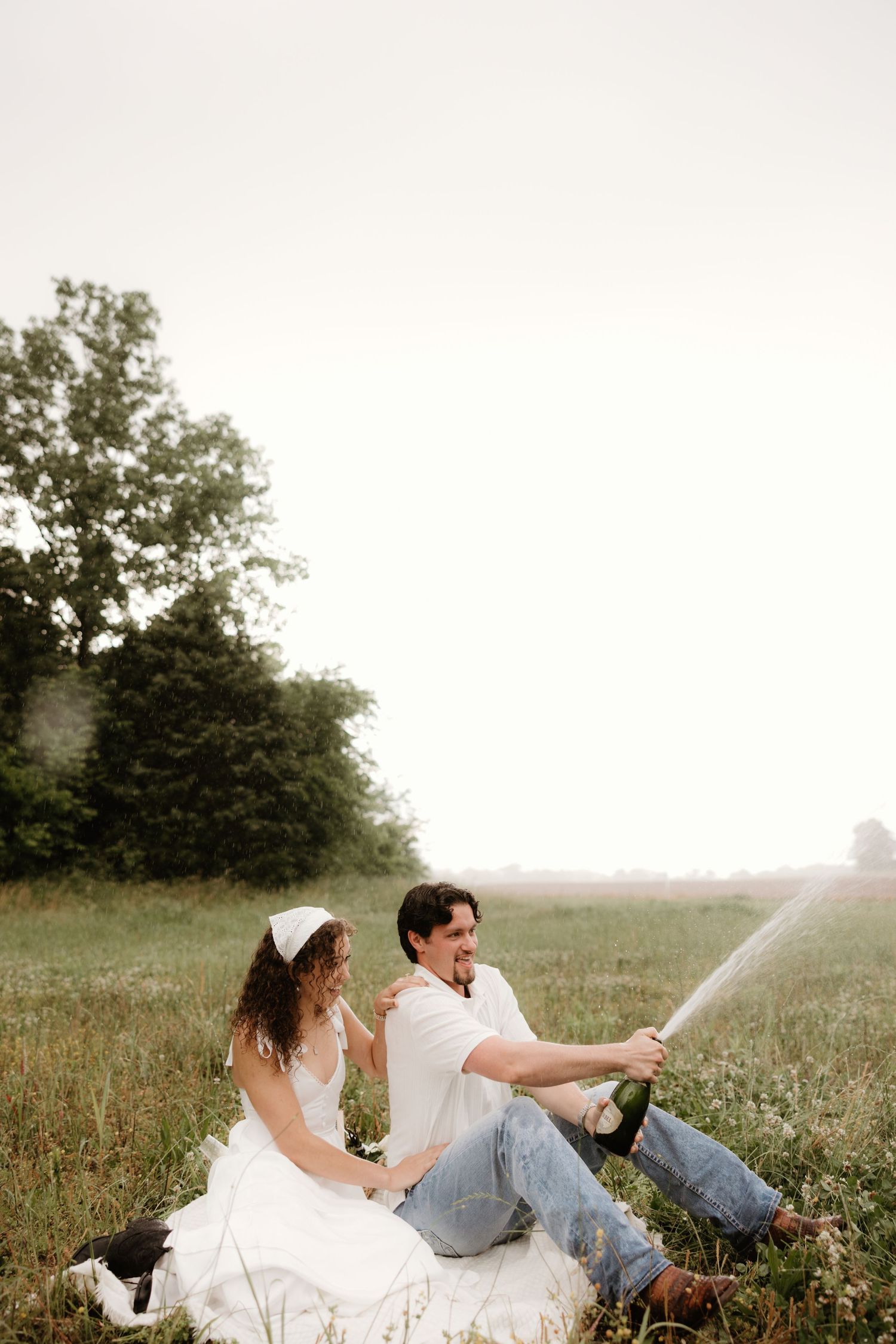 Playful couple sprays champagne while sitting in a meadow.