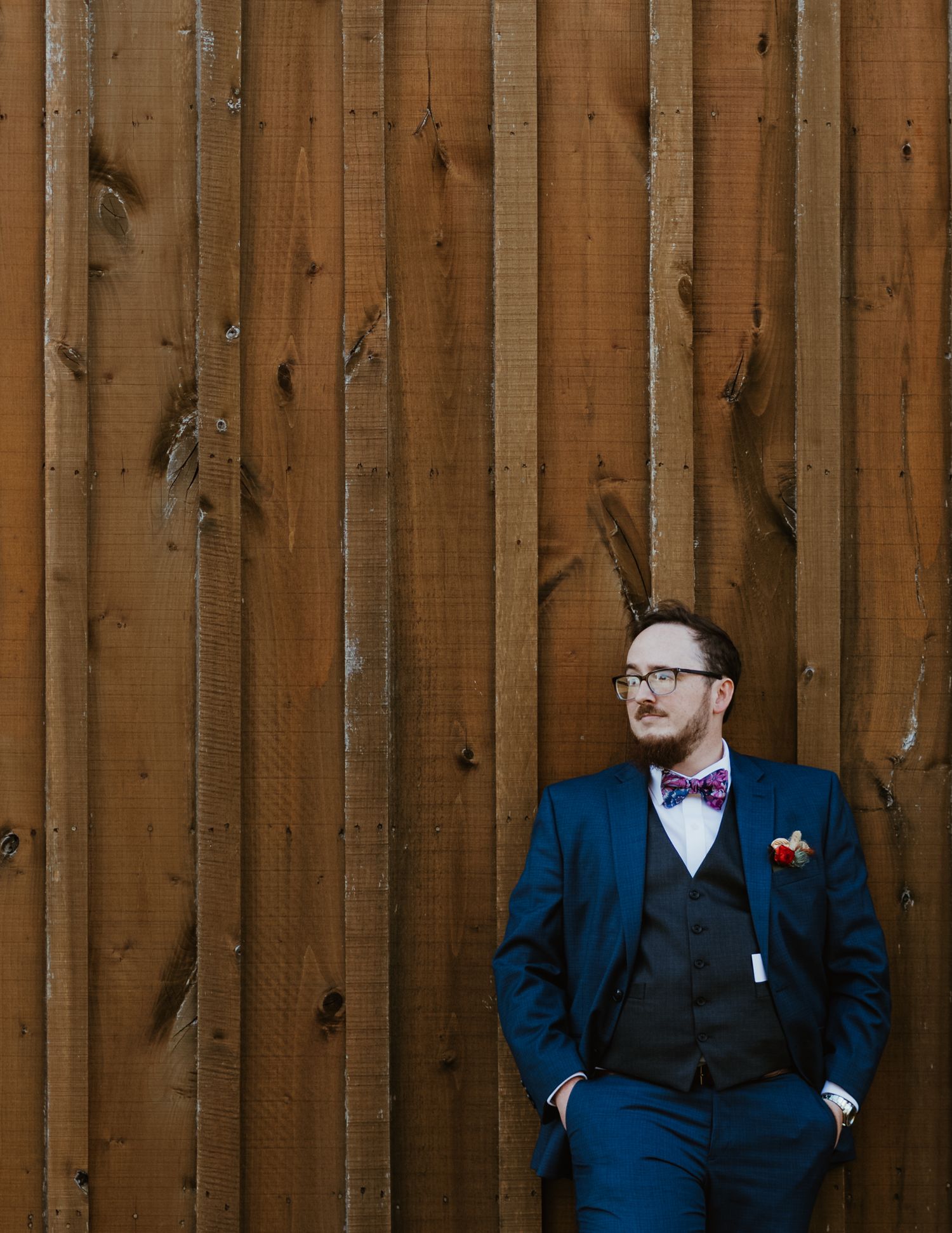 Wedding party members in navy suits posed against rustic wooden fence.
