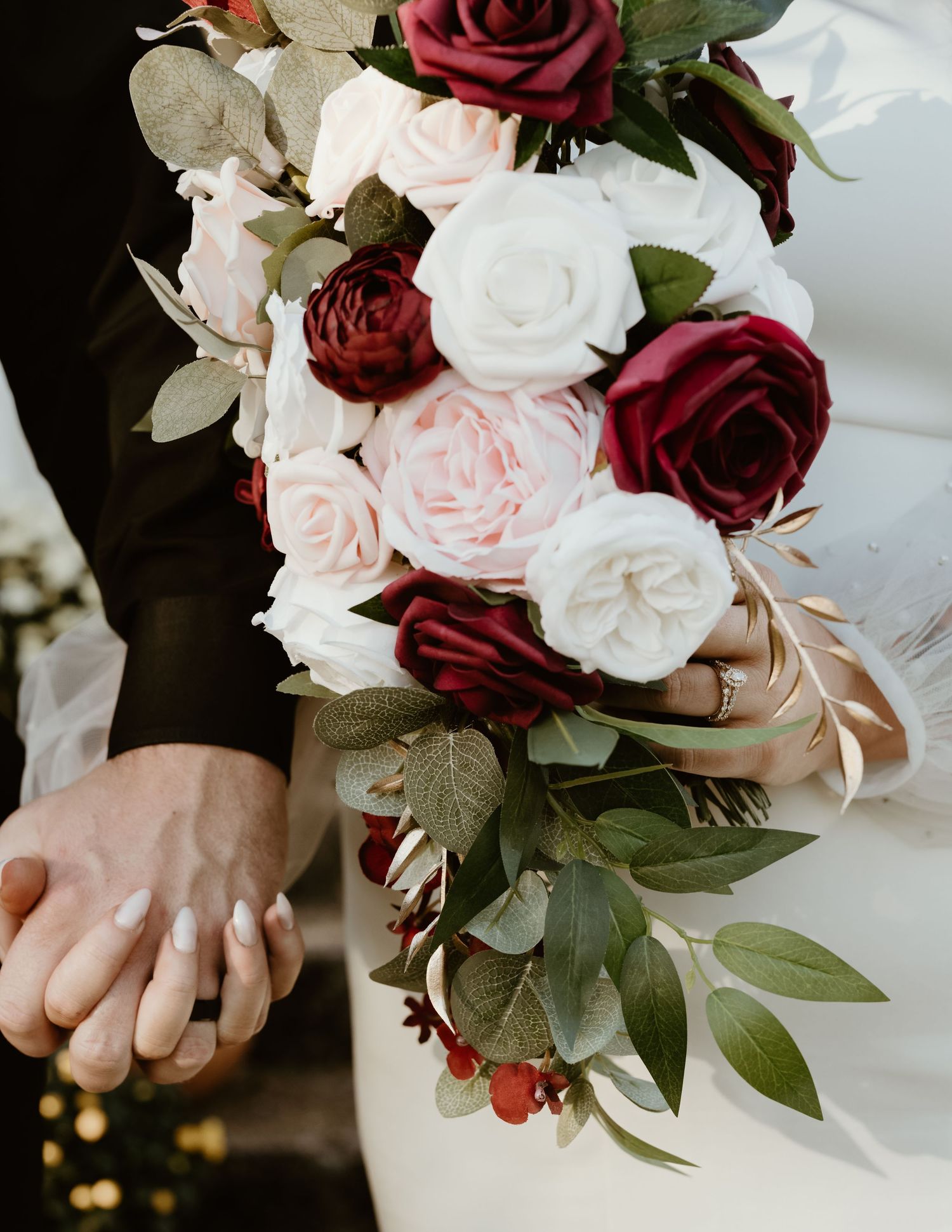 A romantic wedding bouquet featuring deep burgundy and white roses with delicate greenery accents held by gentle hands.