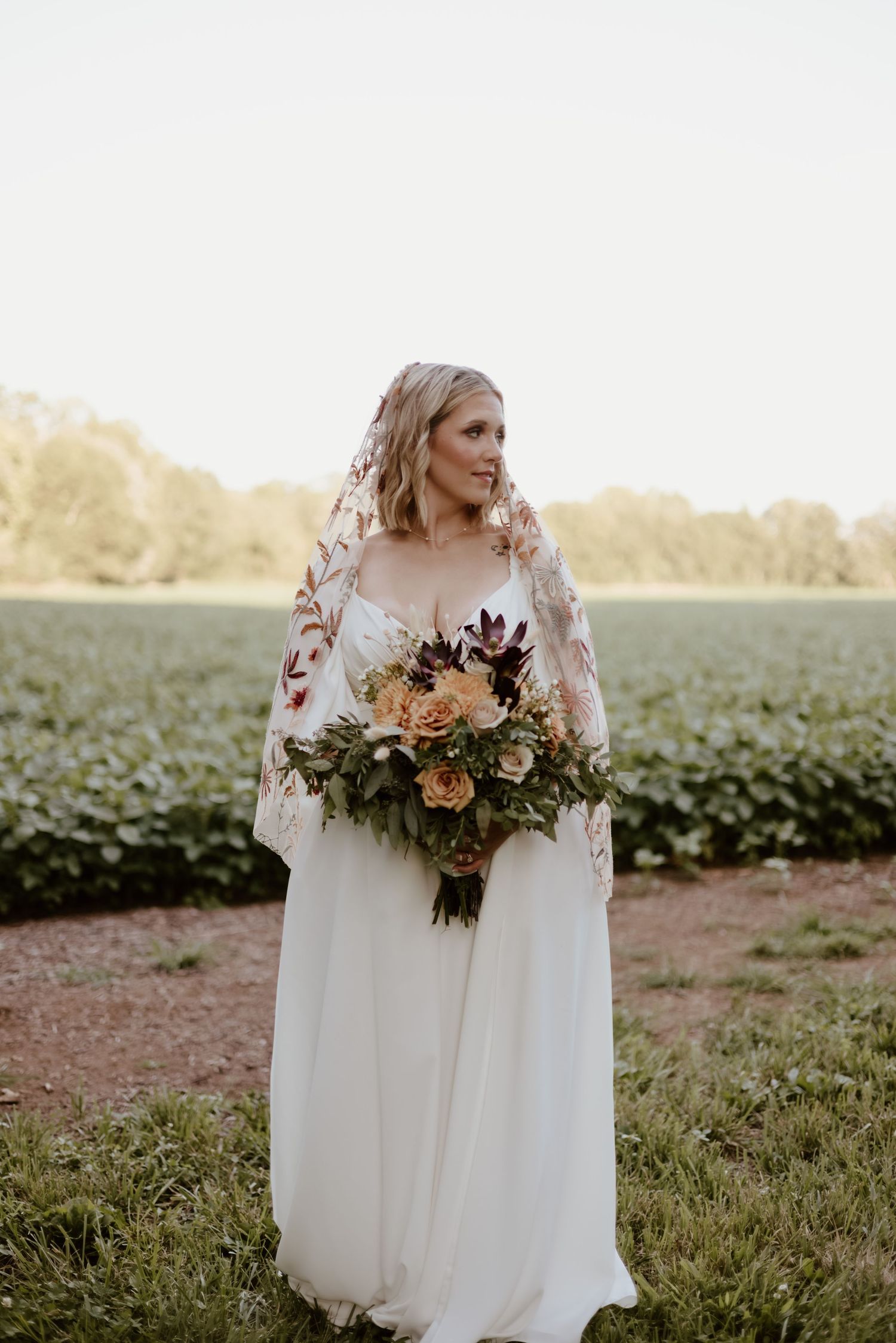 Bride wearing flowing white gown with lace sleeves holds lush autumn bouquet of peach and burgundy flowers in field at sunset.