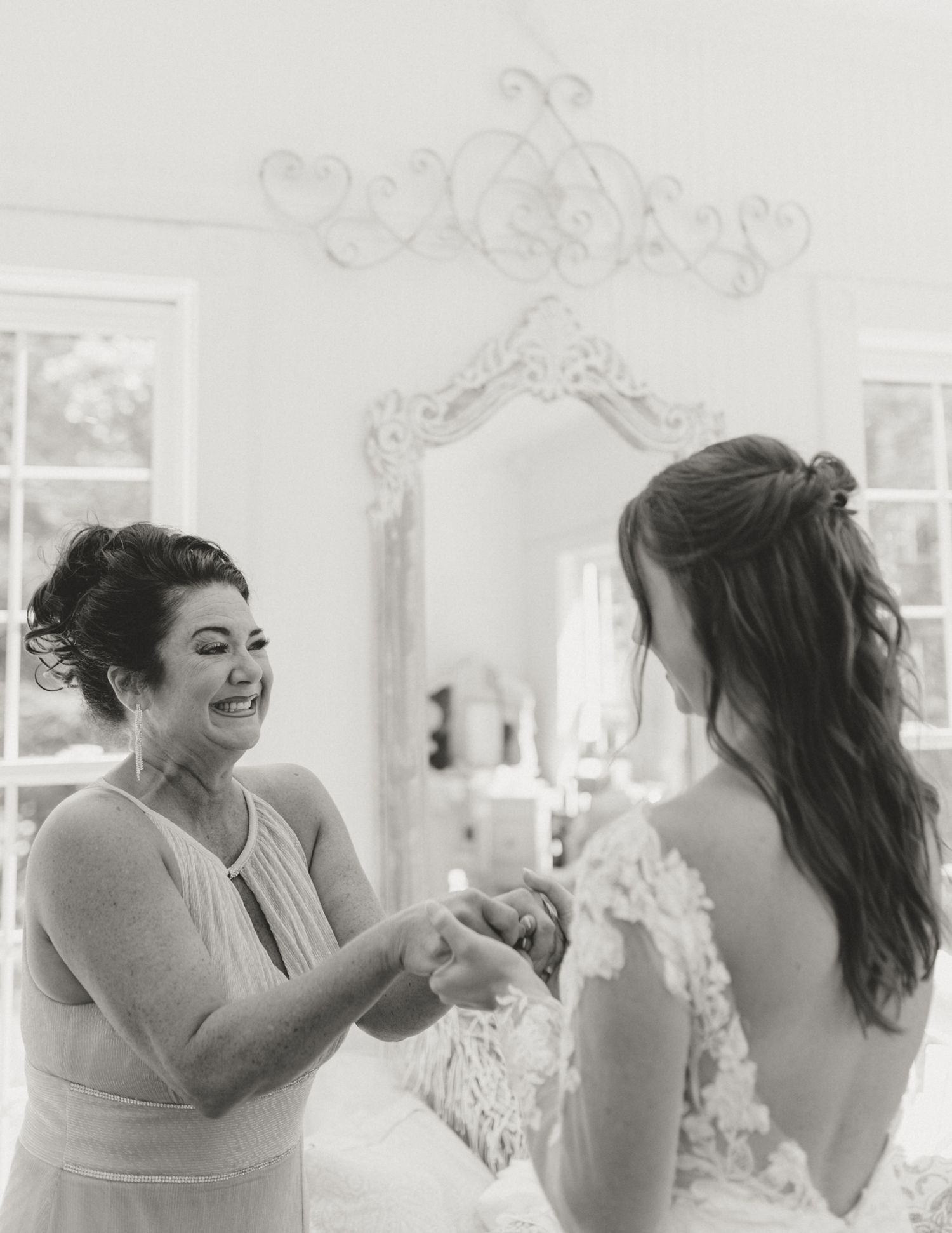 Two women share a joyful moment getting ready in an elegant white room with ornate mirror and decorative moldings.