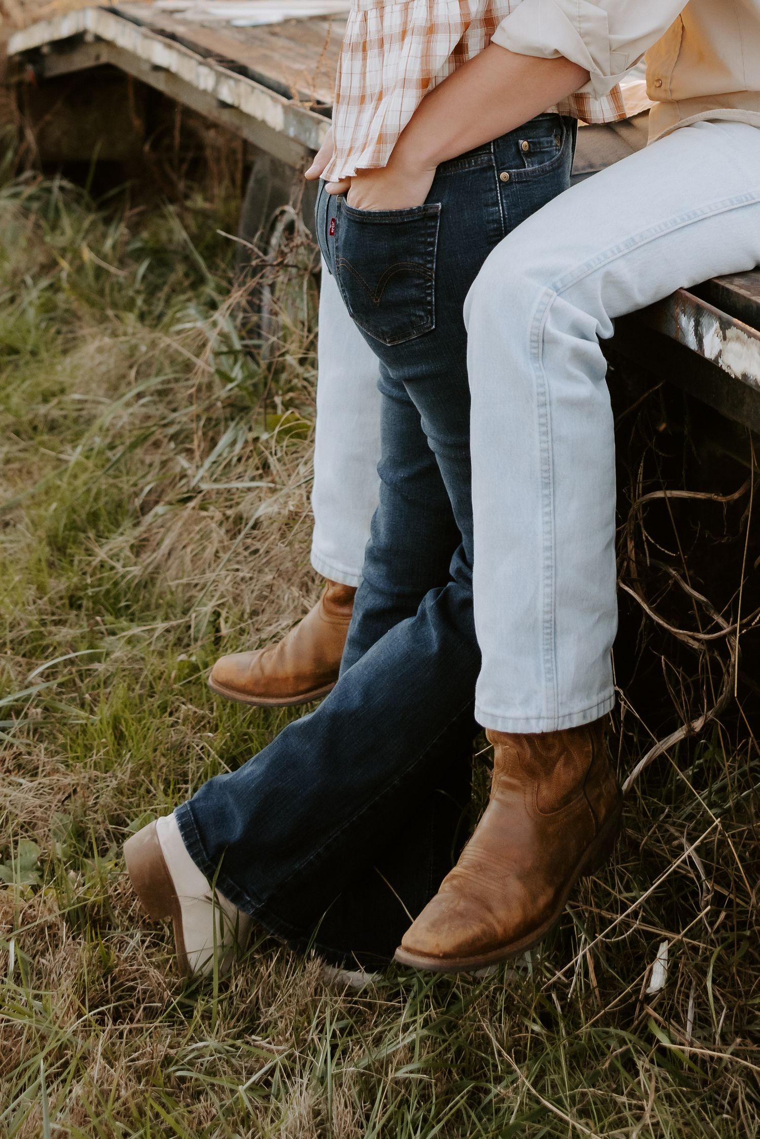 Two people wearing jeans and brown cowboy boots stand in a grassy field near a weathered wooden structure.