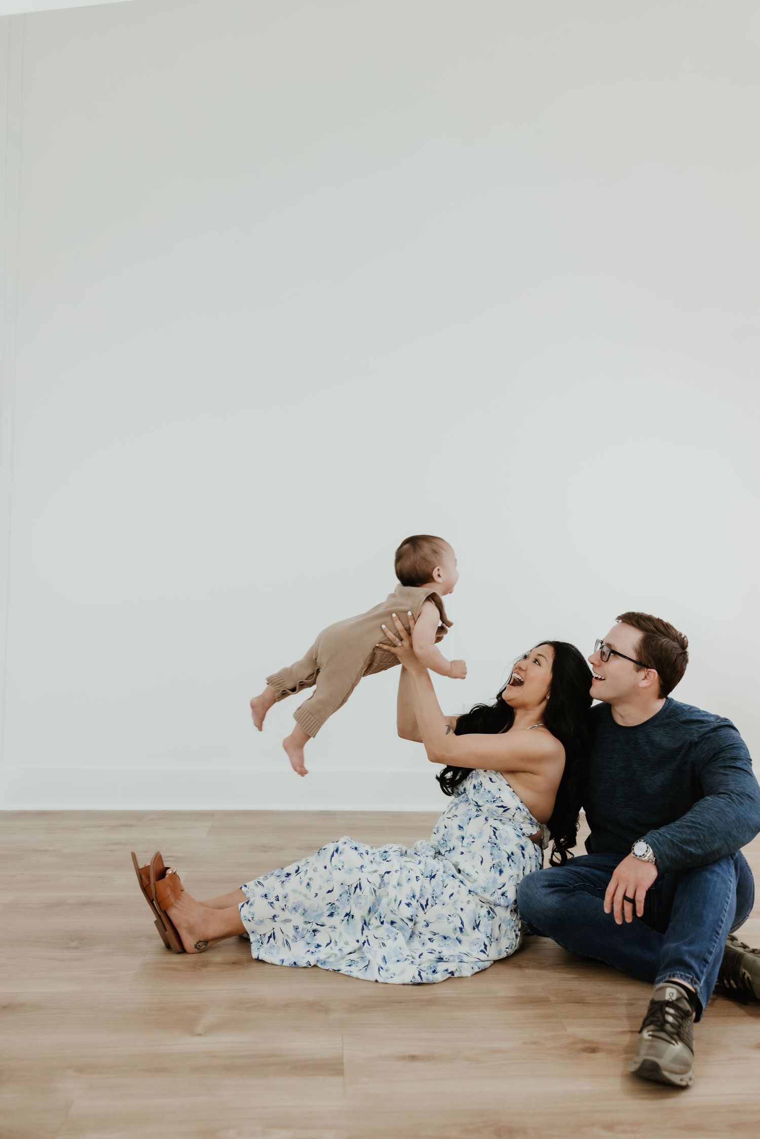 A joyful couple sits on a hardwood floor, the woman lifting a baby in the air while both parents smile lovingly.