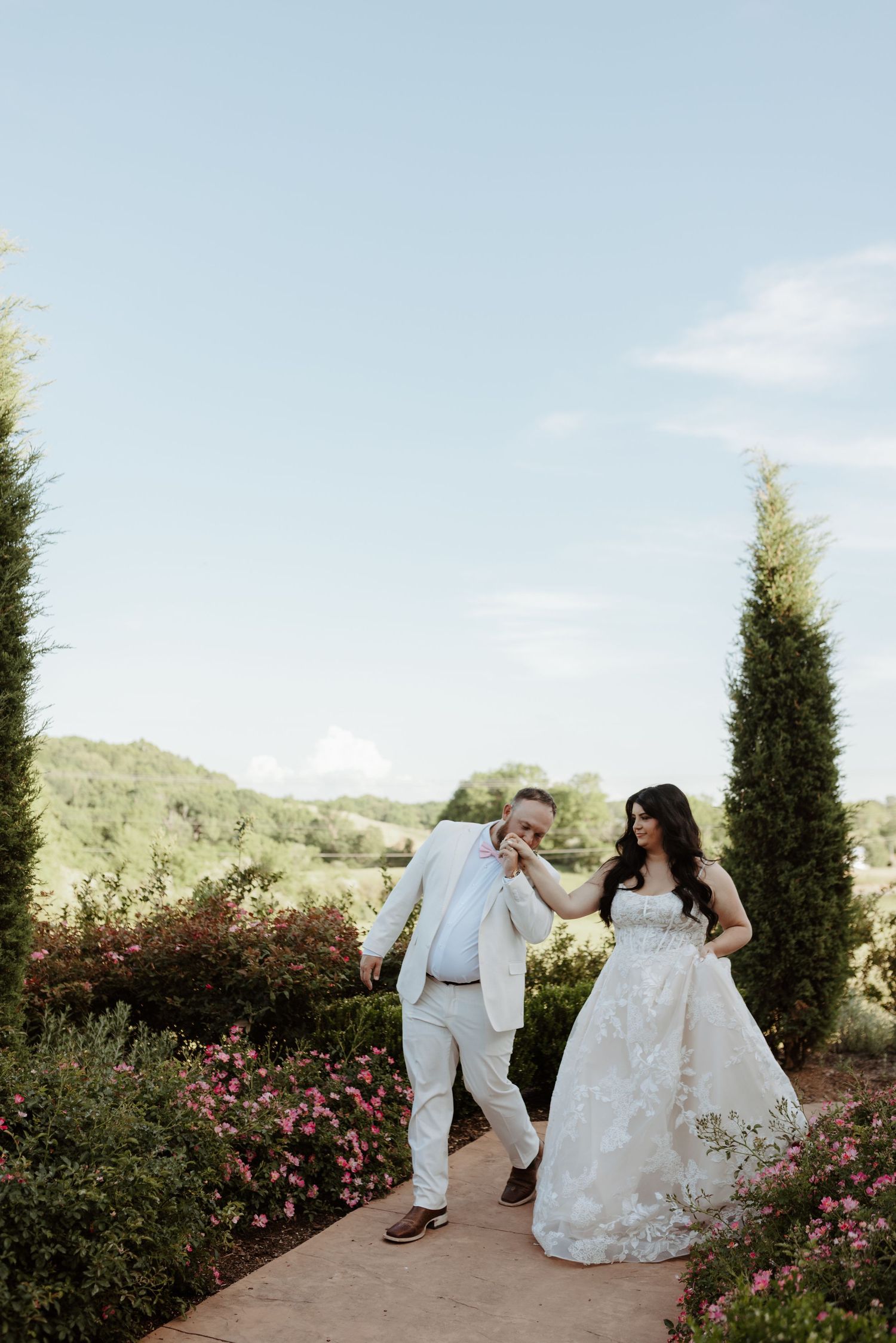 Groom in white suit kisses bride's hand on garden path surrounded by tall cypress trees and pink roses.