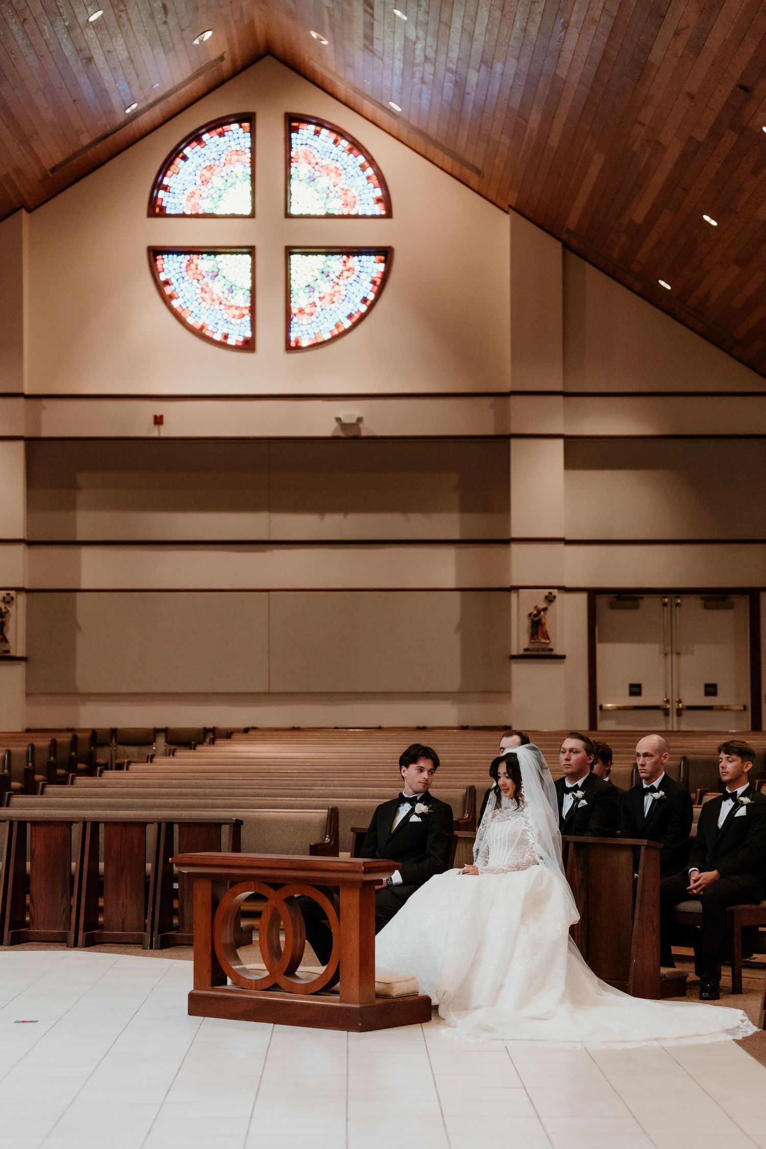 Bride and groom kneel at altar during church wedding ceremony with stained glass rose window backdrop.