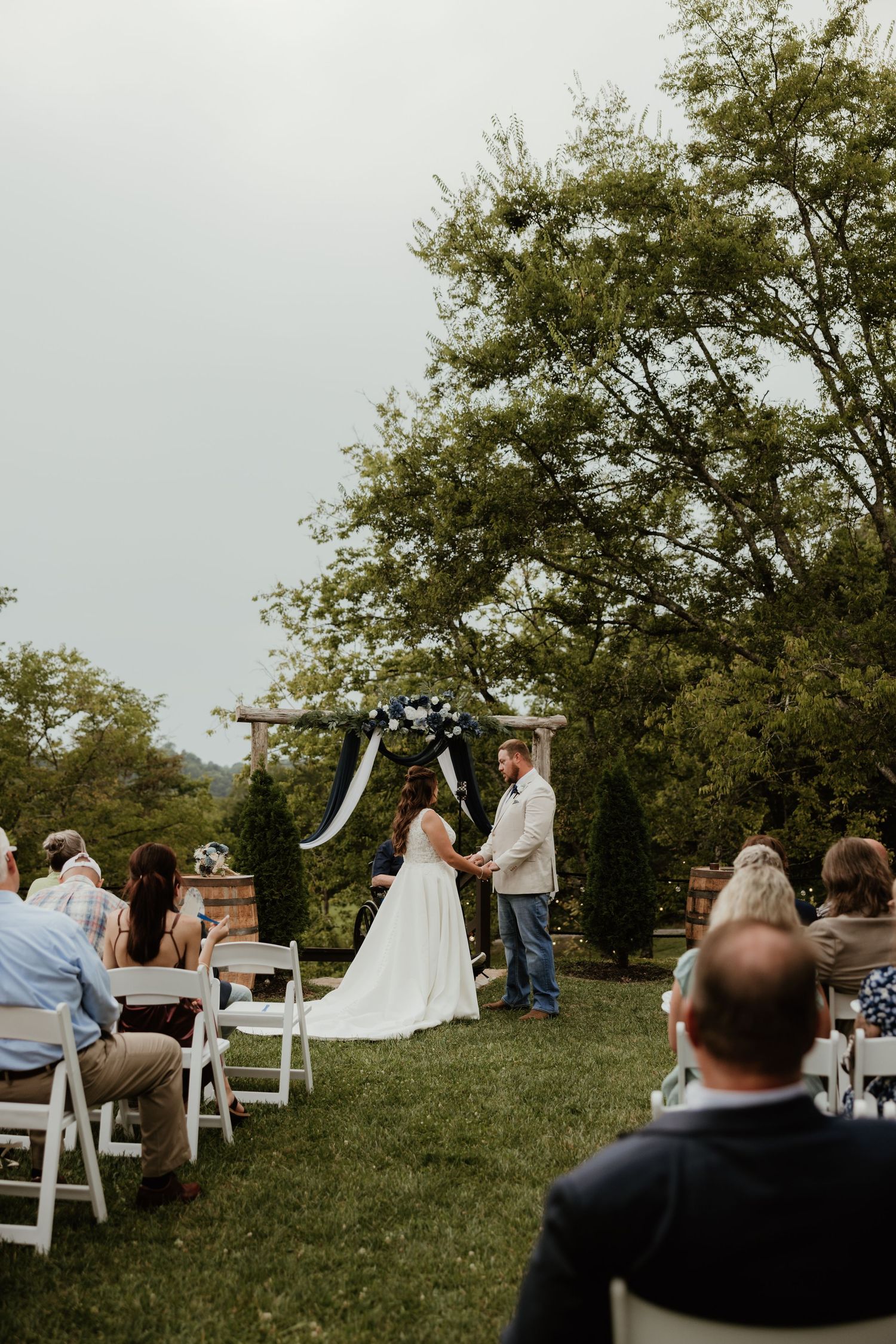 Outdoor wedding ceremony takes place on grassy hillside with white chairs and trees in background.