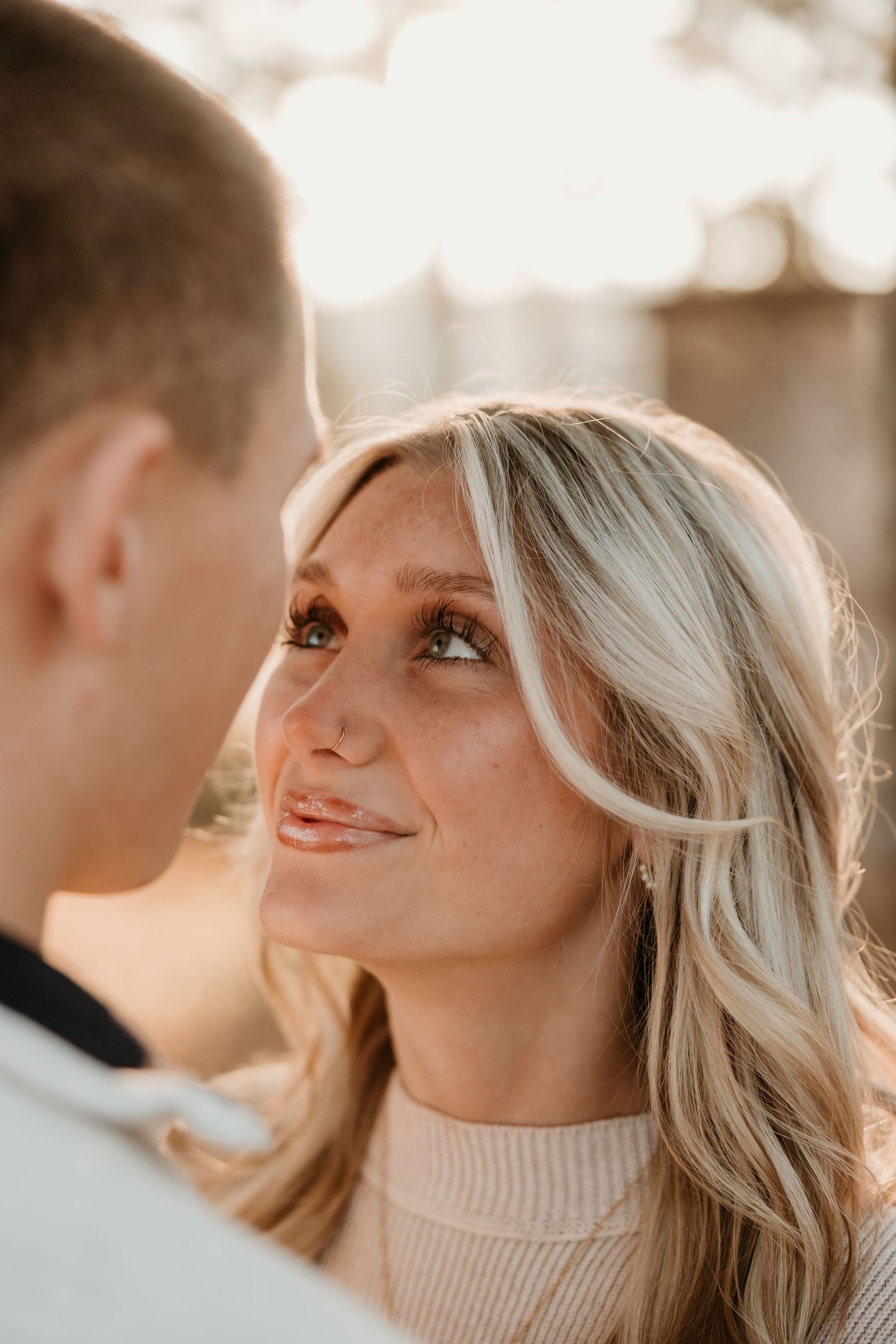 A blonde woman gazes lovingly at a man in a warm golden light during an outdoor couples engagement photo session.