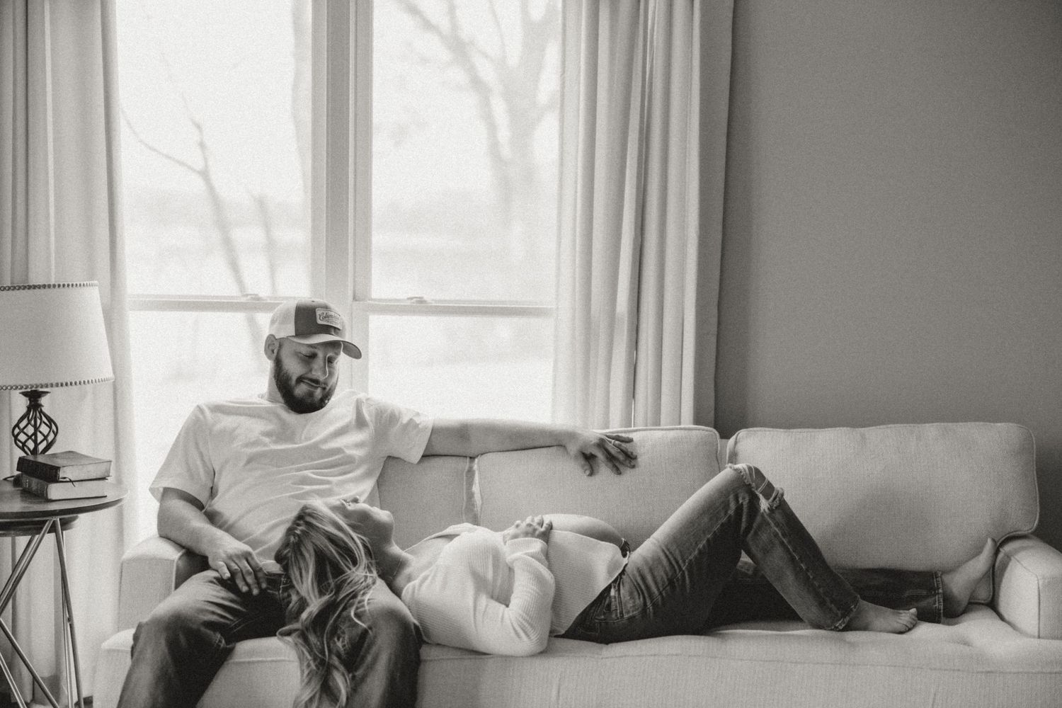 Black and white photo of a couple relaxing on a couch near a window, man in cap, woman resting her head on his lap.