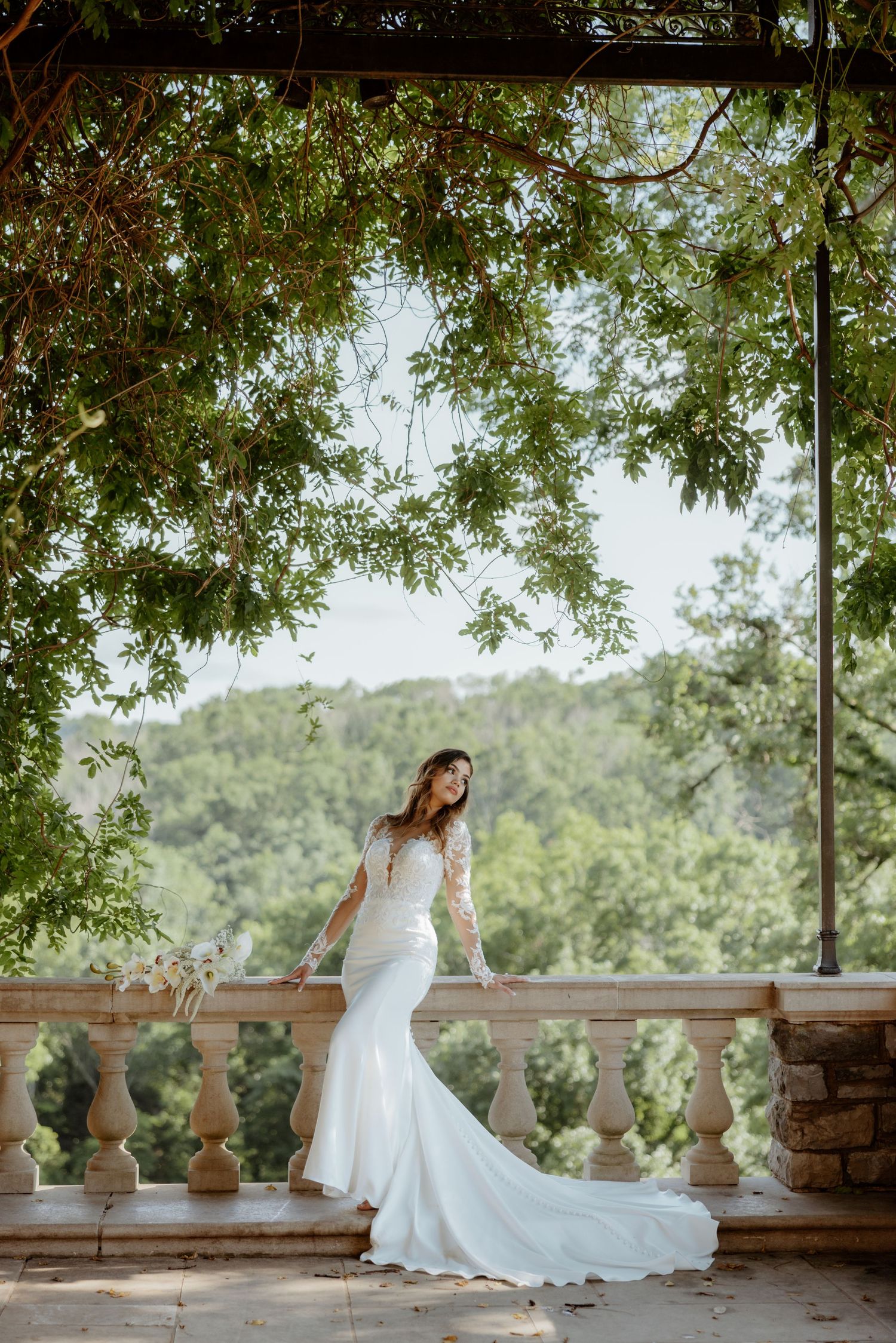 Bride in lace mermaid gown poses on stone balcony under lush green canopy with rolling hills backdrop.