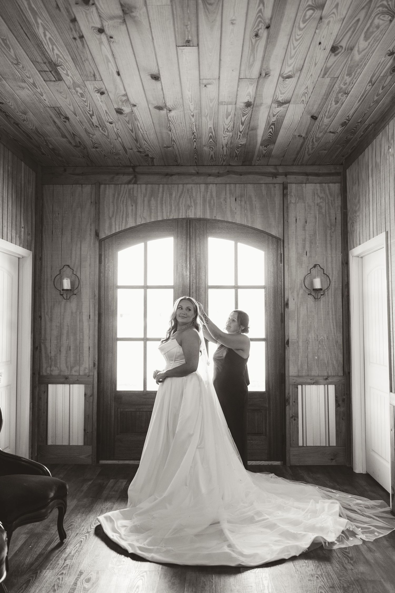 A black and white wedding photograph showing a long white dress with a dramatic train in a rustic wooden room.