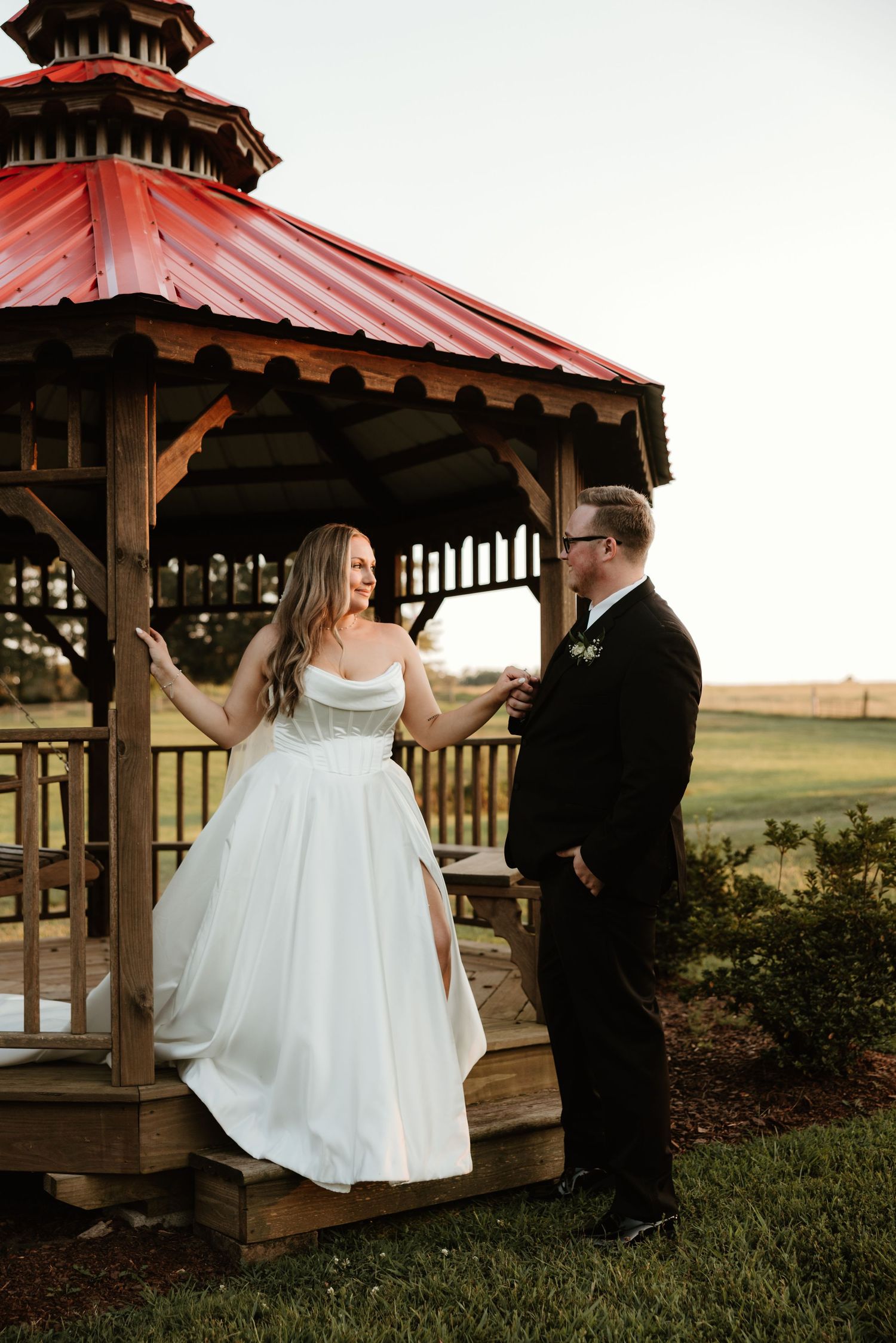 A romantic wedding moment captured in a rustic gazebo with red roof during sunset.
