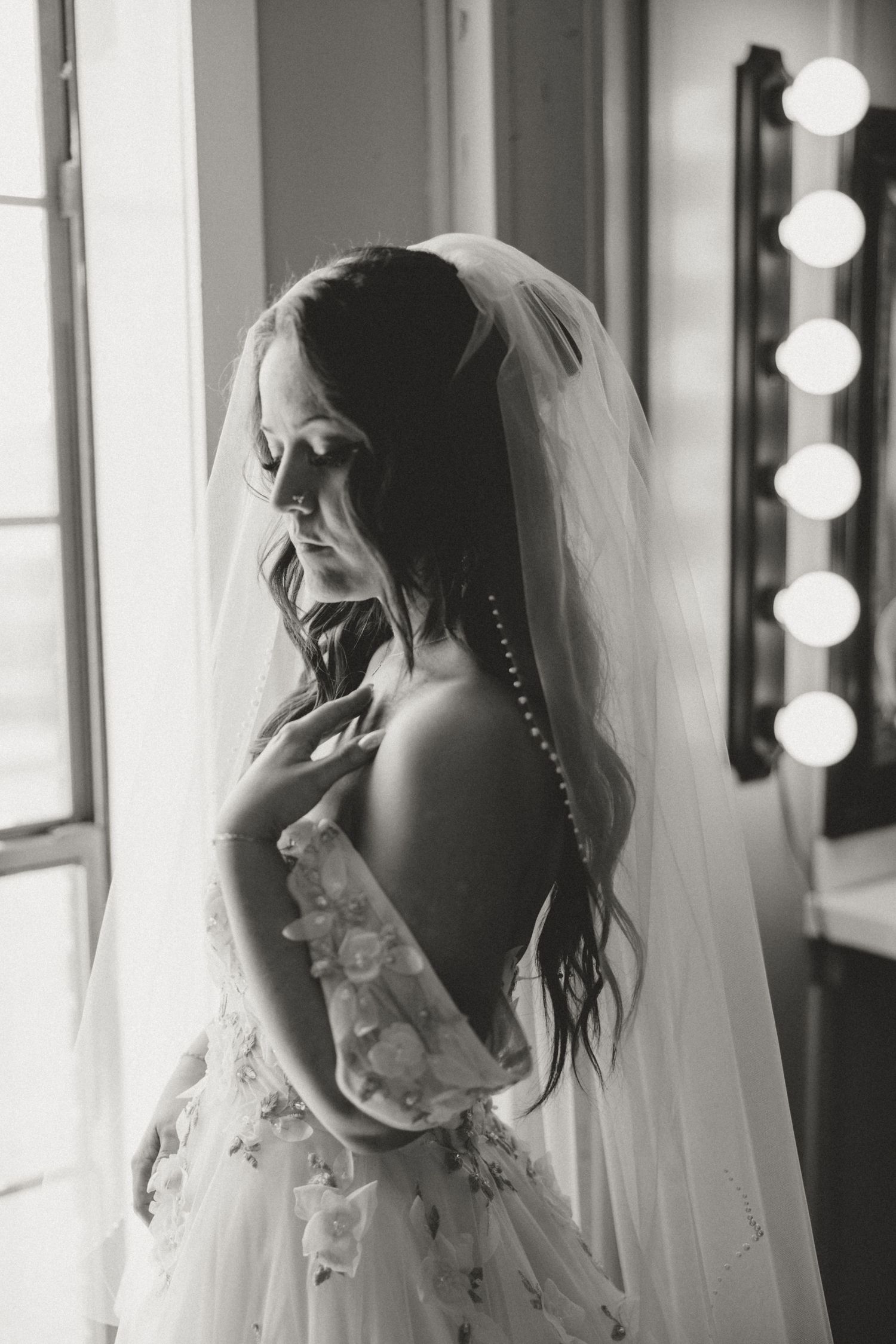 A bride wearing a delicate veil and white gown poses near a window with illuminated makeup mirrors in black and white.