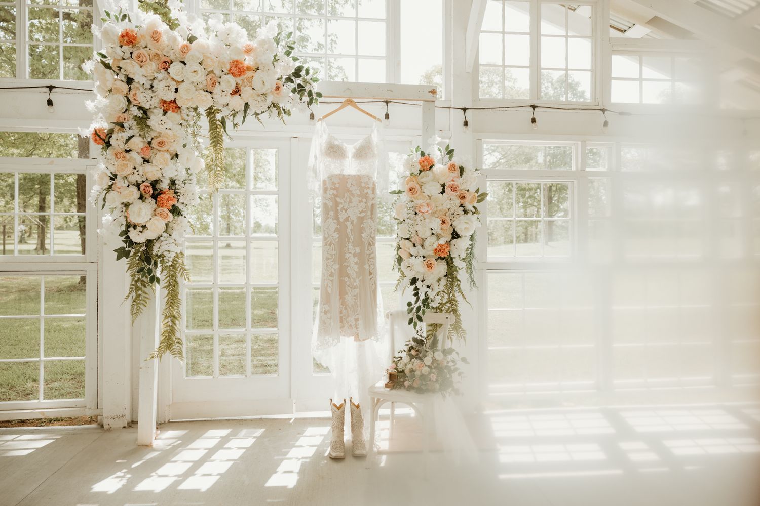 A dreamy wedding dress hangs between two floral arch arrangements in a sunlit white conservatory space.