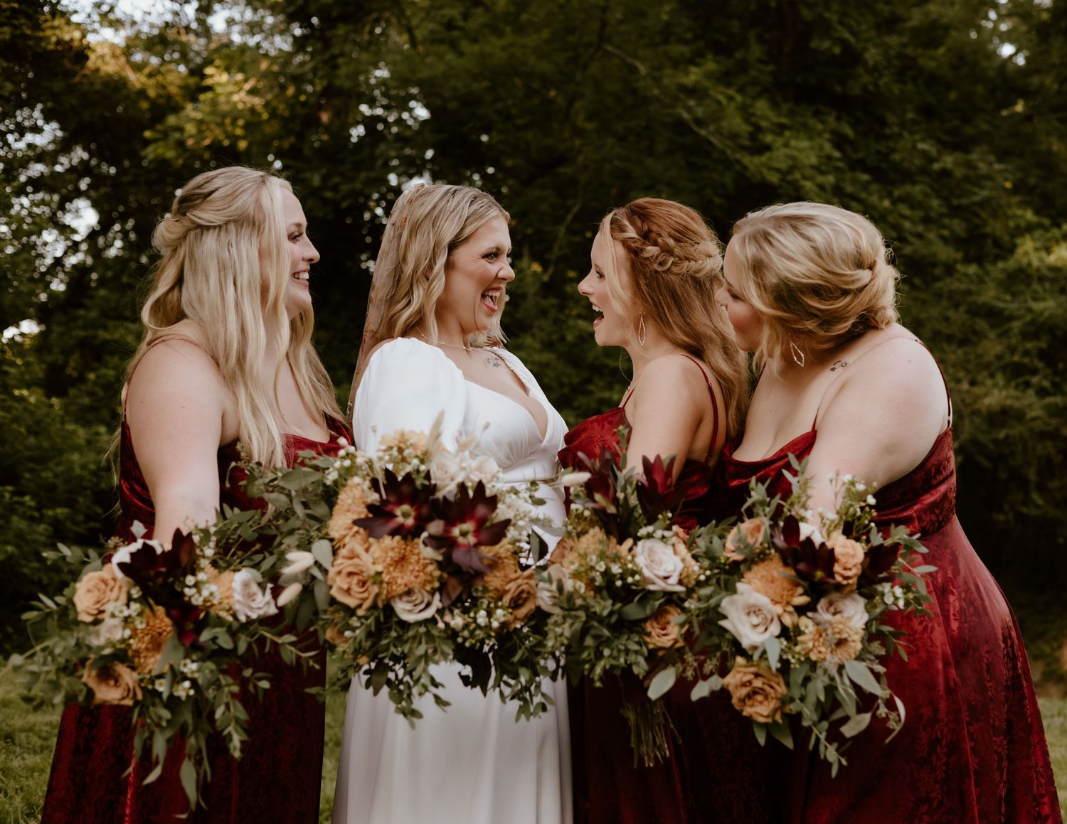 Bridesmaids in burgundy dresses and bride in white hold autumn-colored bouquets while standing outdoors.