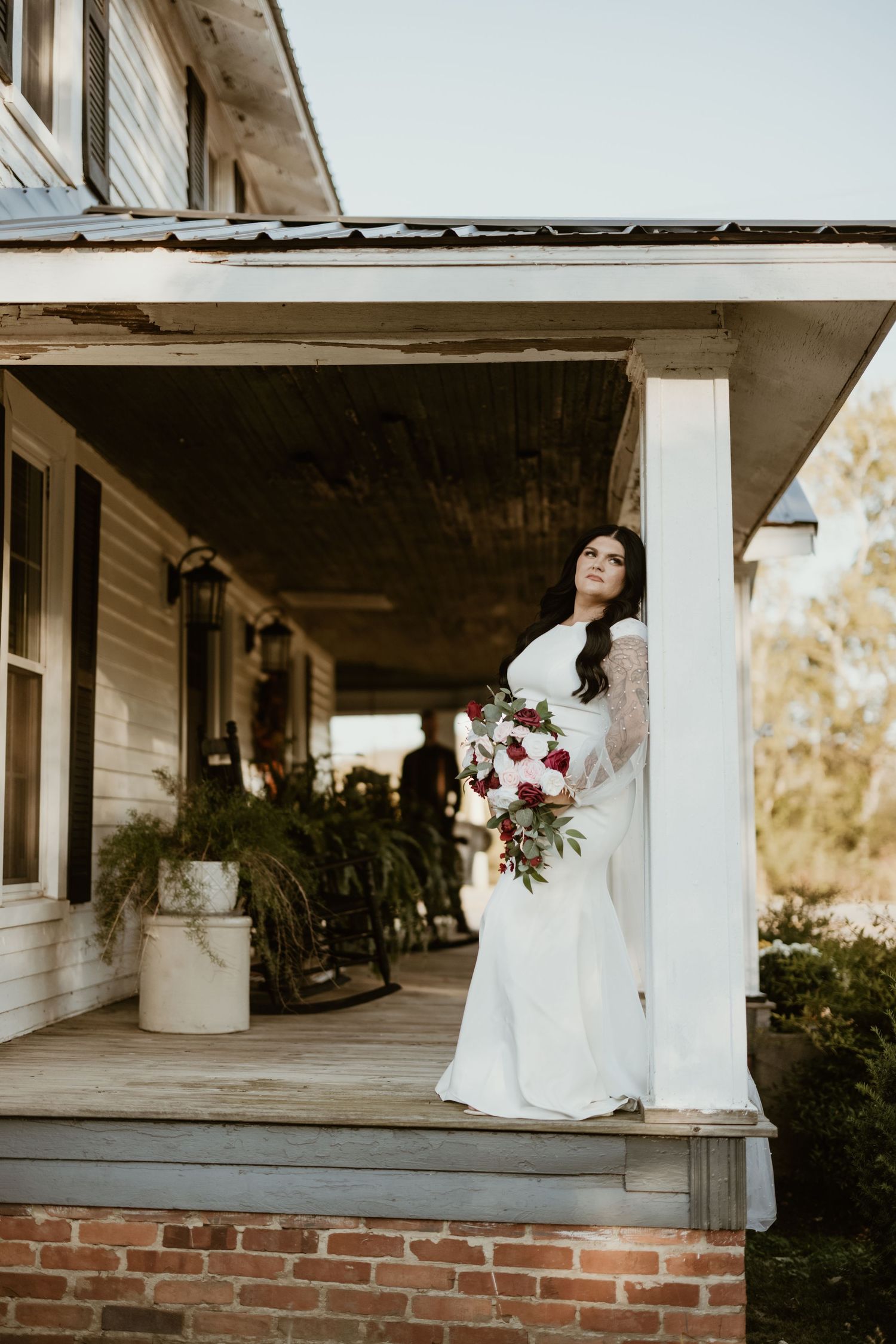 A bride in a white lace gown holds a deep red bouquet while standing on a rustic farmhouse porch at sunset.