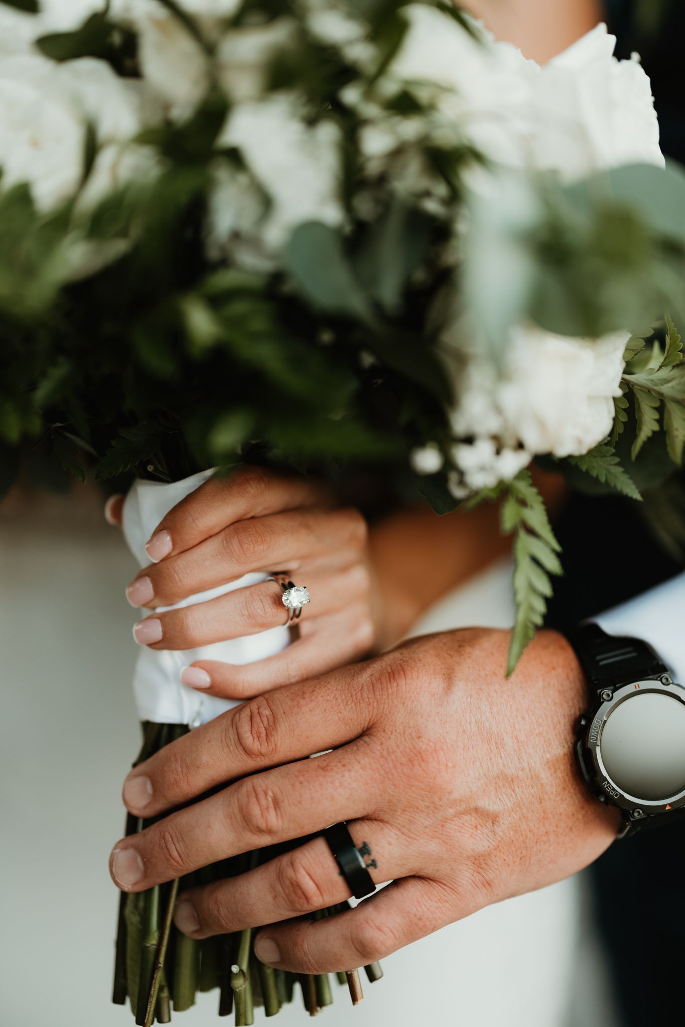 Close-up of hands wearing wedding rings while holding white flowers and greenery against a blurred background.