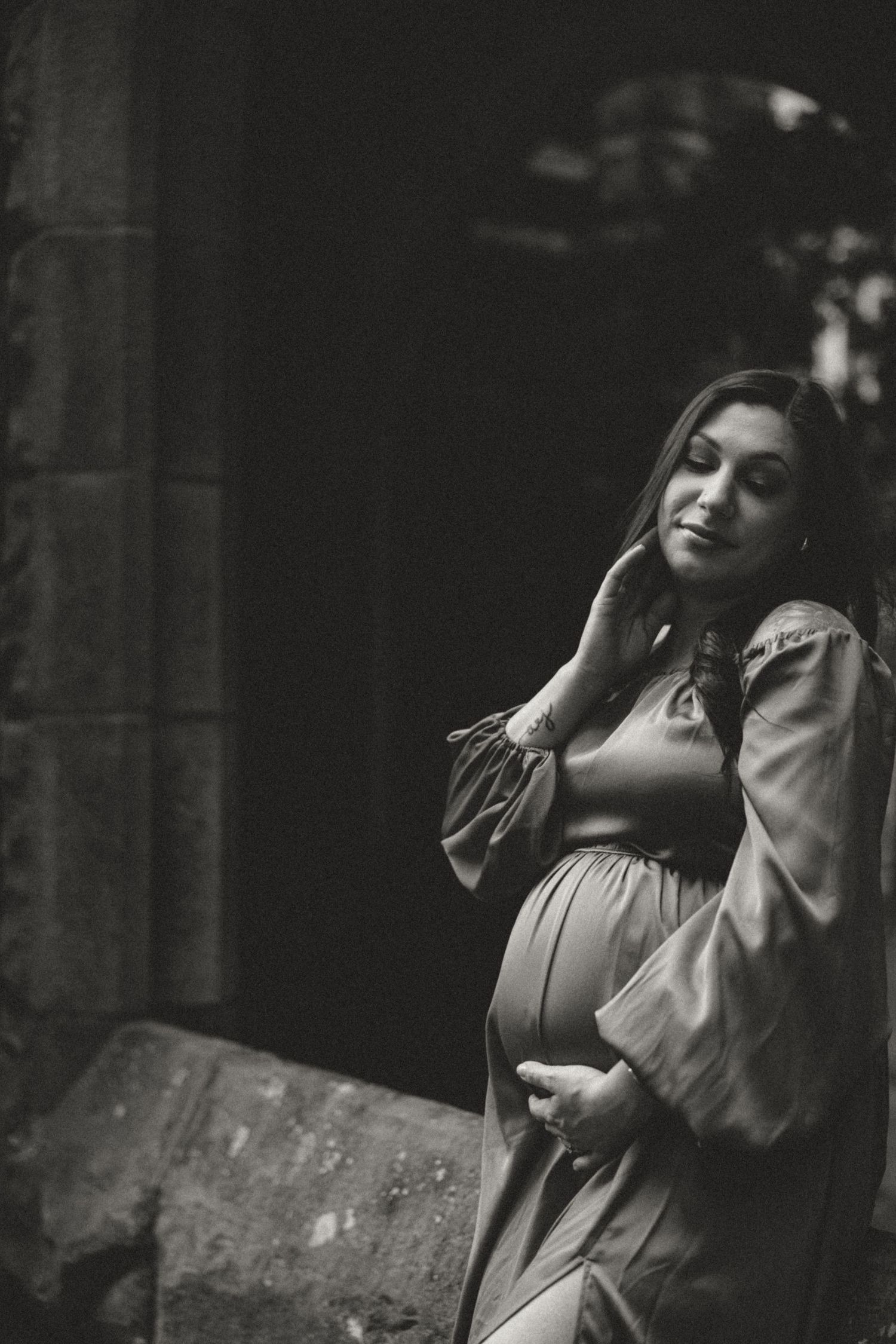 A black and white maternity portrait featuring dramatic lighting and stone architecture.