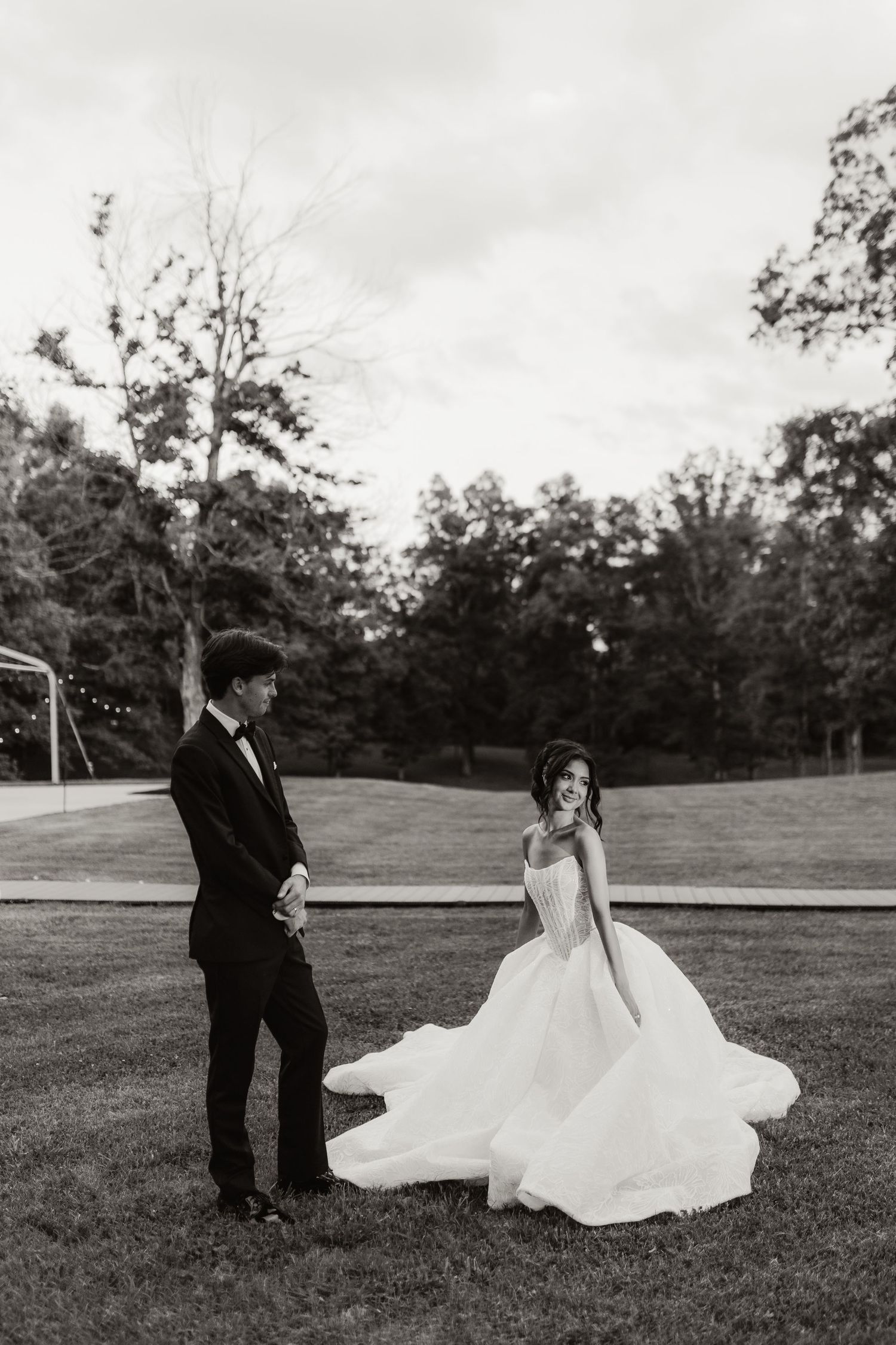 Black and white photo of bride in flowing ball gown and groom in tuxedo standing on green lawn.