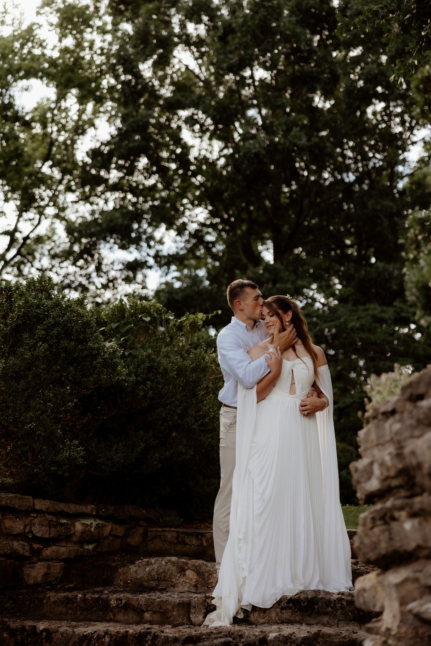 Couple in white wedding attire embracing on stone steps surrounded by lush green trees in an outdoor ceremony.