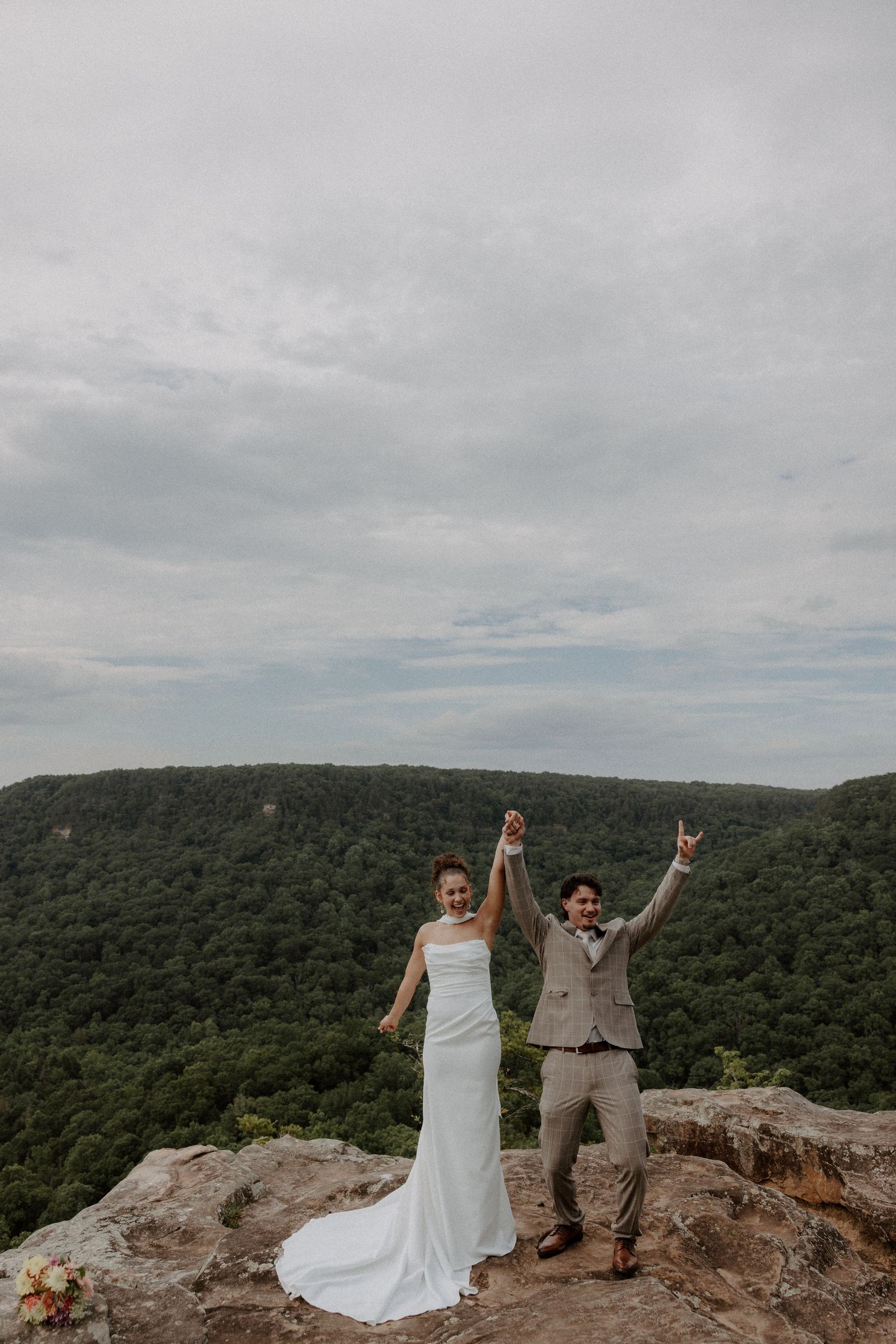 Bride and groom celebrate on a rocky mountain overlook with arms raised, wearing wedding attire against a lush forest backdrop.