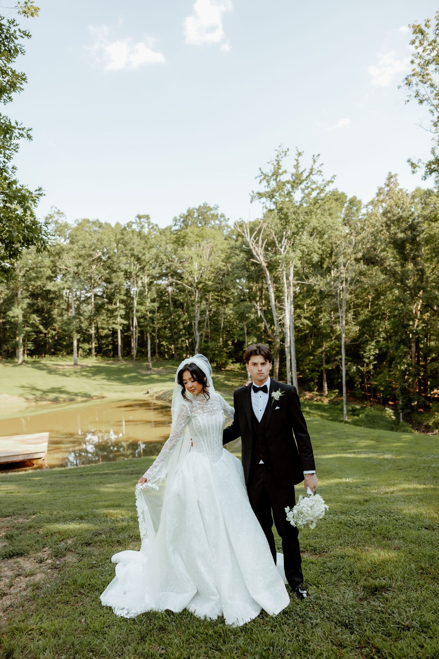 Bride and groom in elegant wedding attire walk hand in hand on green lawn with forest and pond backdrop.