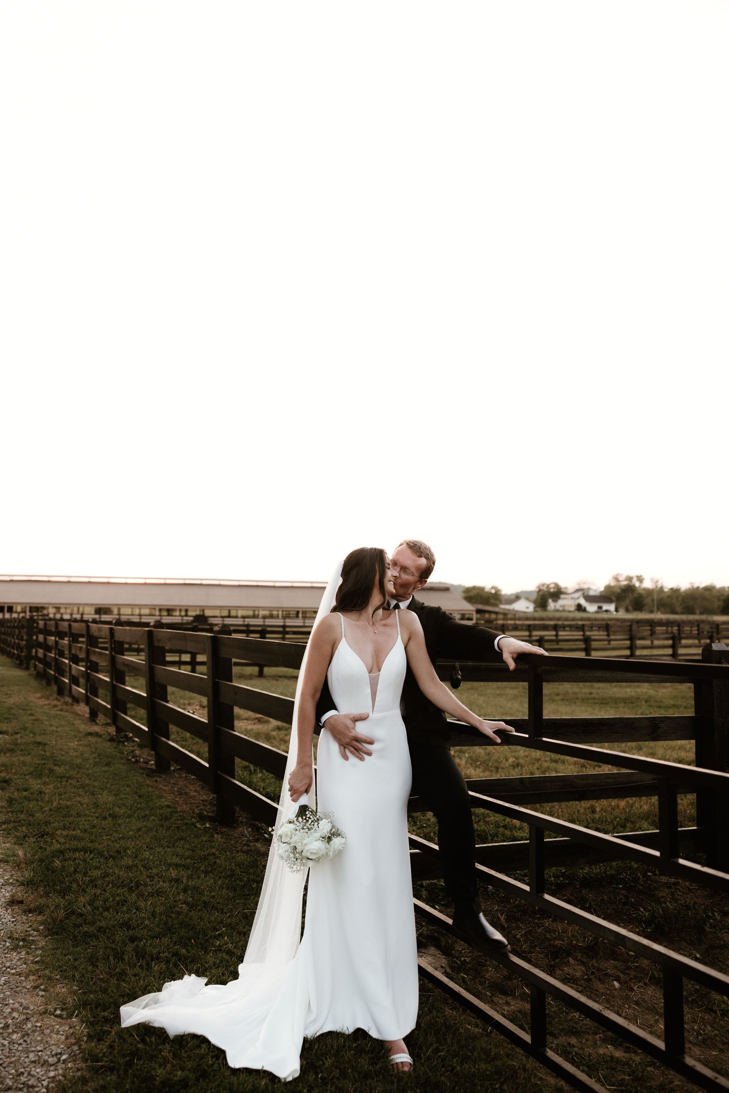 Elegant wedding couple standing by a wooden fence in a rustic countryside setting.