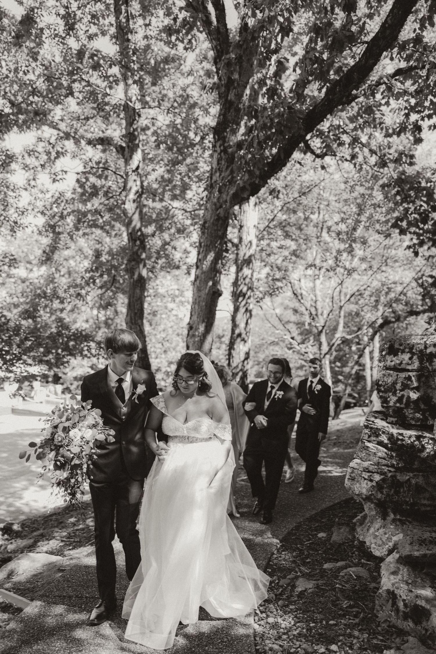 A bride and groom walk through a scenic wooded area surrounded by wedding guests on a sunny day.