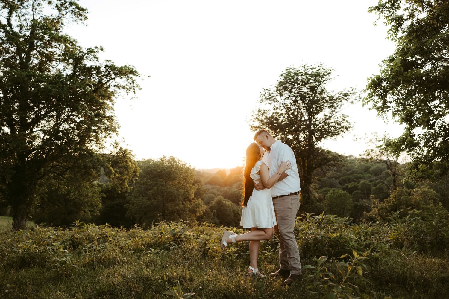A couple in white embraces during golden hour sunset in a scenic hilltop meadow surrounded by trees.
