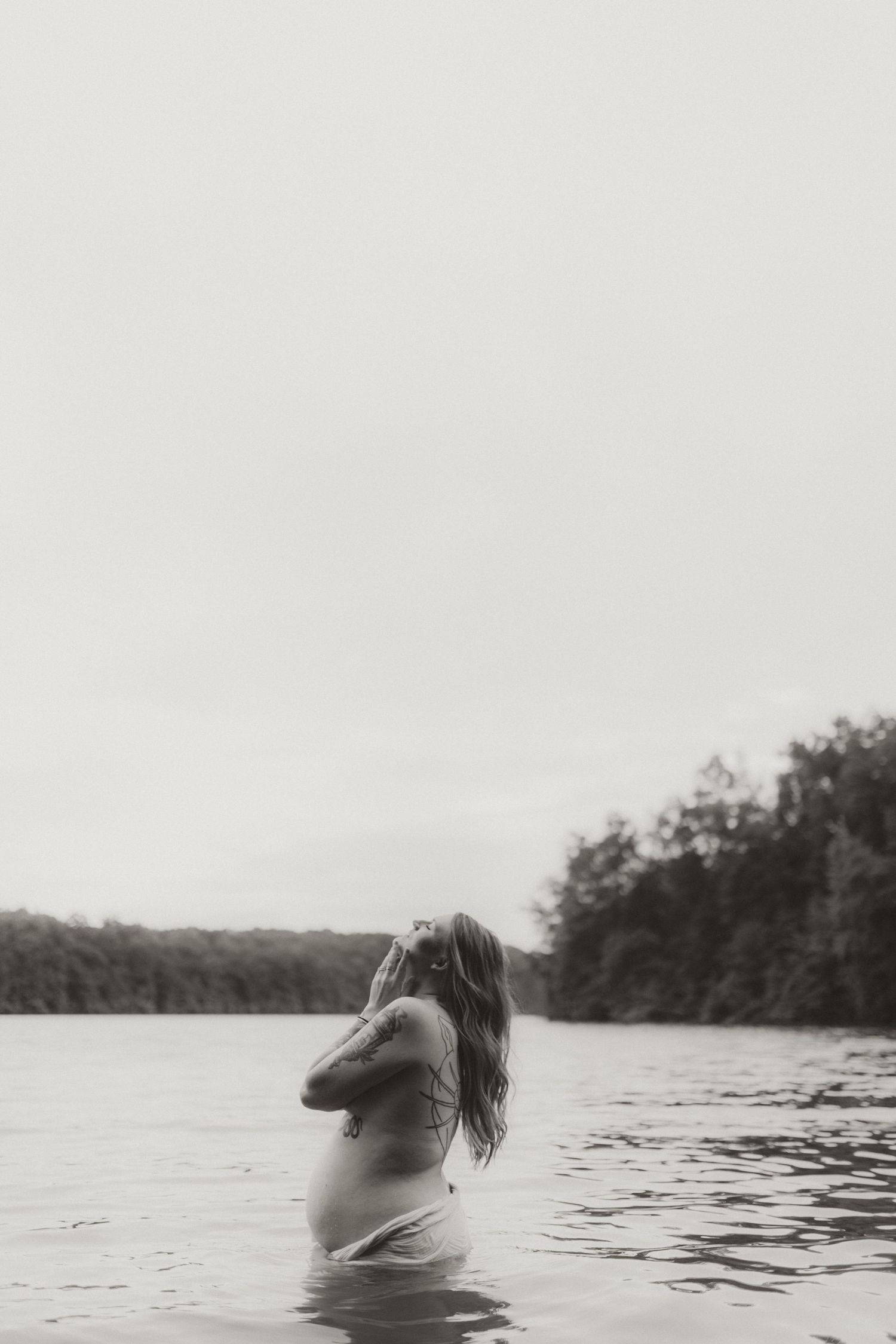 A person stands waist-deep in a serene lake surrounded by trees during a tranquil black and white moment.
