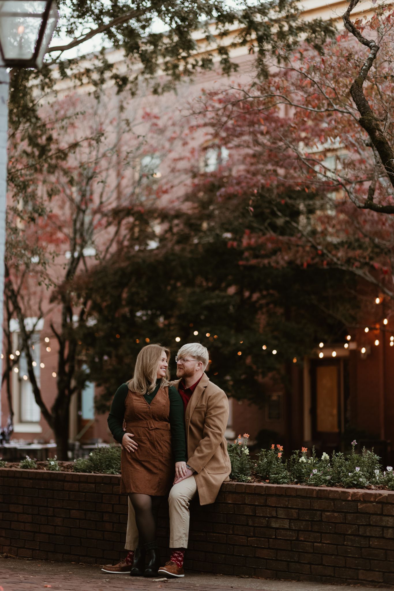 A couple sits together on a brick wall under blooming trees with string lights creating a romantic autumn atmosphere.