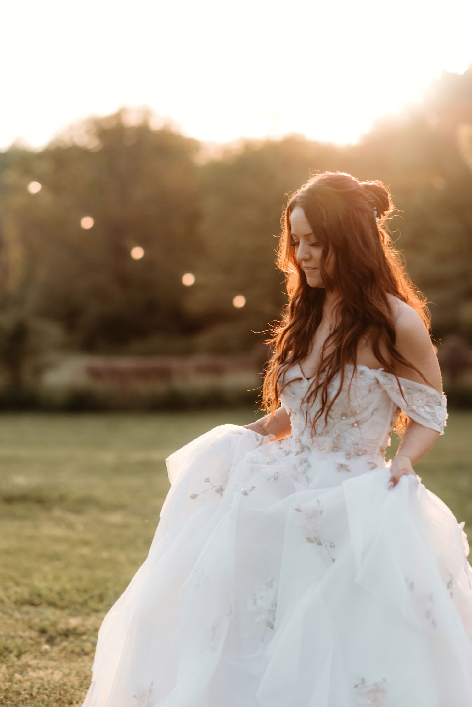 A flowing white wedding dress catches golden sunlight in a grassy field at sunset.