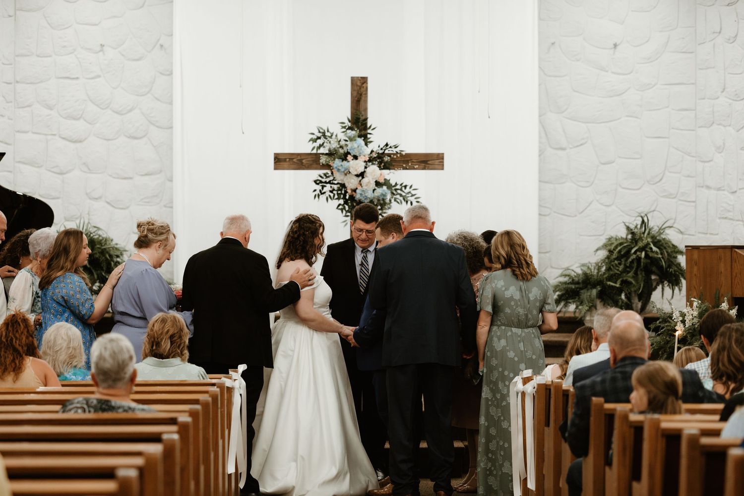 A wedding ceremony takes place at the altar of a white-walled church with wooden pews and a simple wooden cross.
