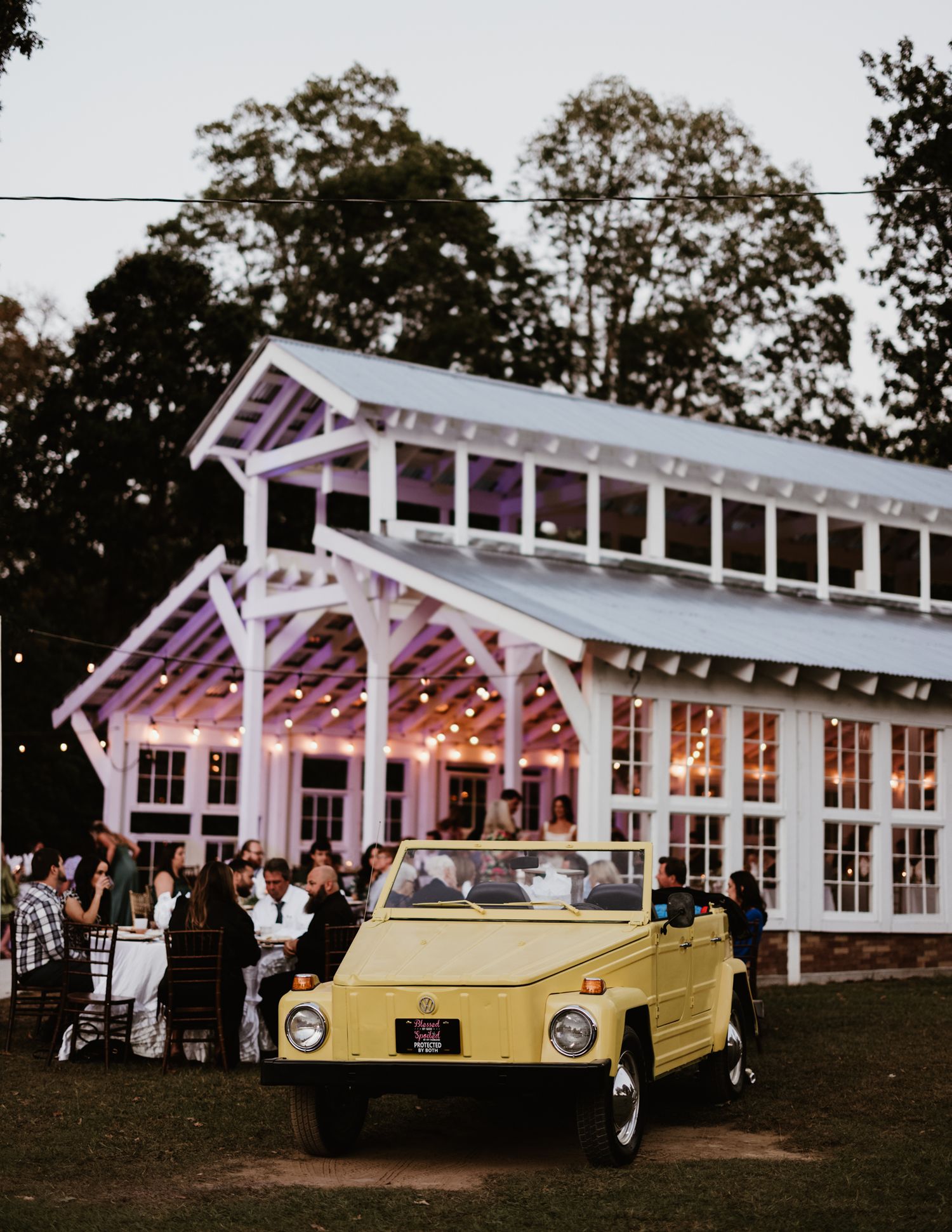 Yellow vintage car parked in front of a white greenhouse-style wedding venue illuminated with string lights at dusk.