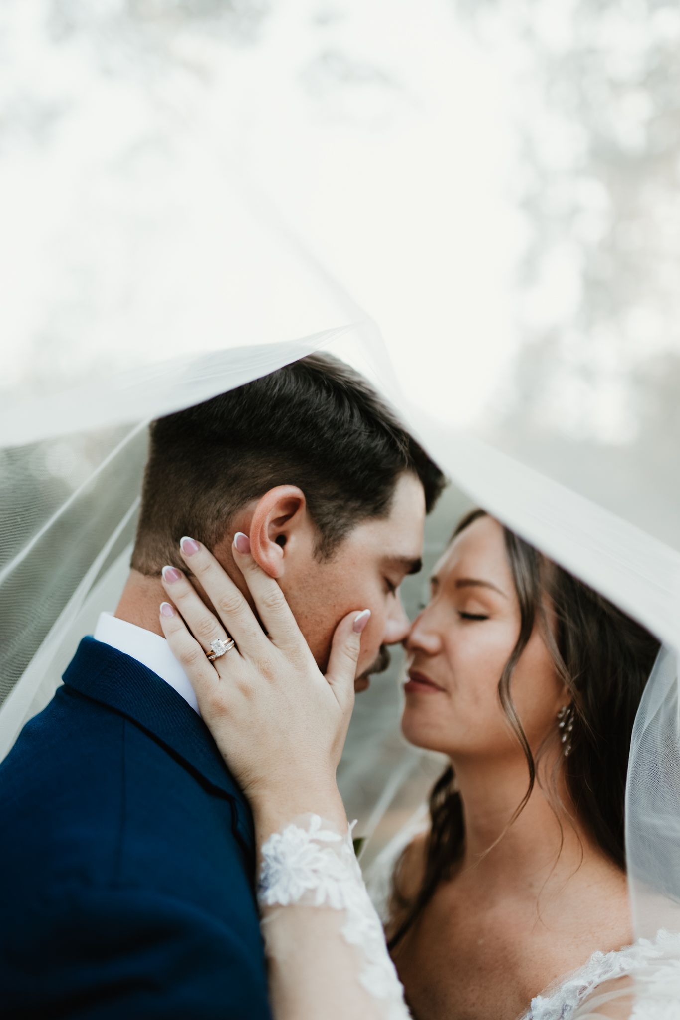 Romantic wedding couple shares an intimate kiss under a delicate veil in soft natural lighting.