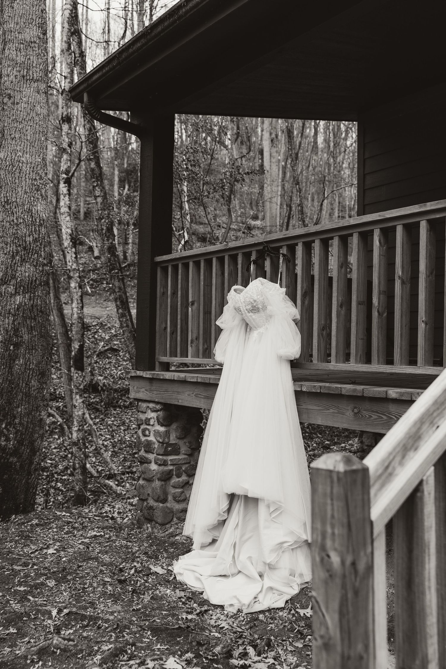 Black and white photo of a bride in a flowing wedding gown and veil standing on a rustic wooden porch surrounded by trees.