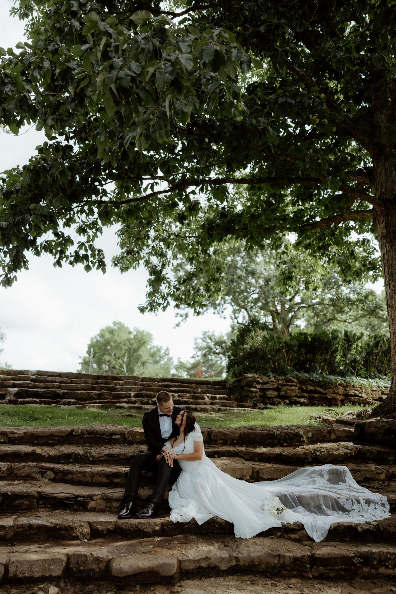 Bride and groom sit together on stone steps under a large tree, bride's veil flowing gracefully in the breeze.