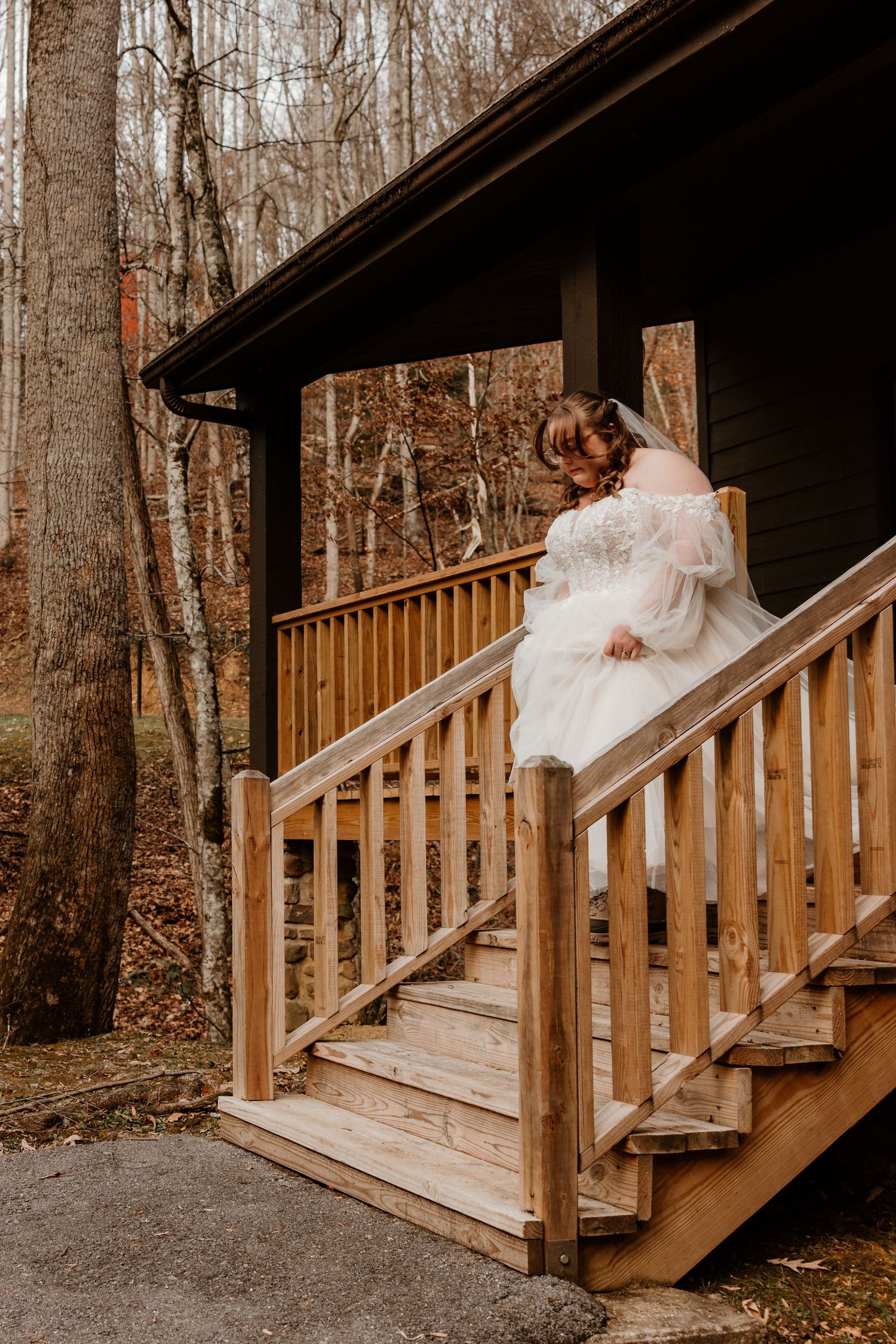 Bride in white lace gown descending wooden cabin stairs surrounded by autumn forest trees.
