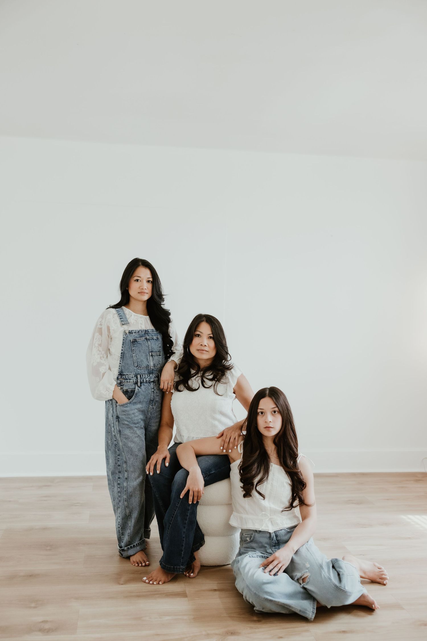 Three young Asian women pose casually in a bright white studio, wearing denim and white tops on a light wood floor.