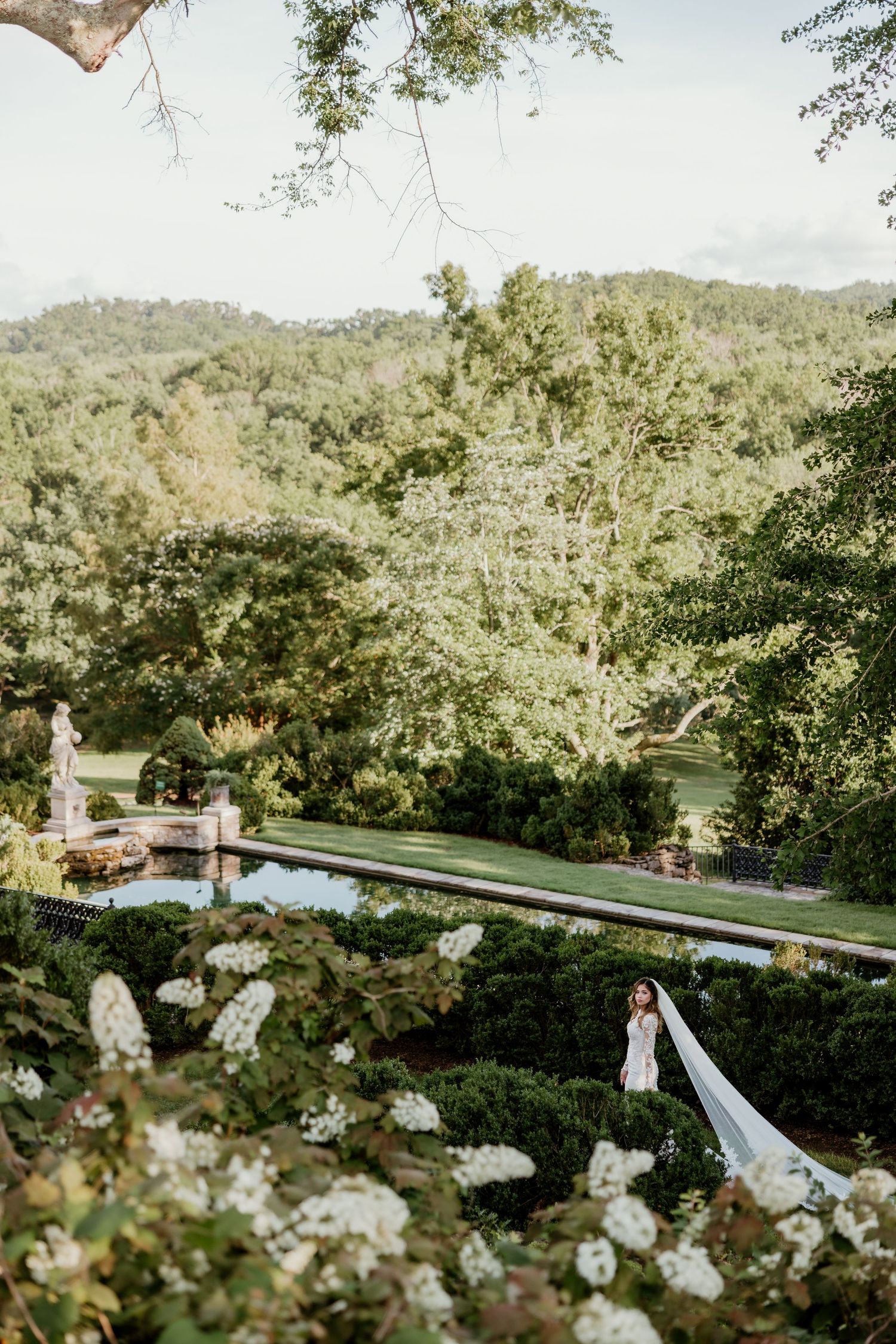 Bride in white gown with long veil stands in lush garden estate with reflecting pool and rolling green hills.