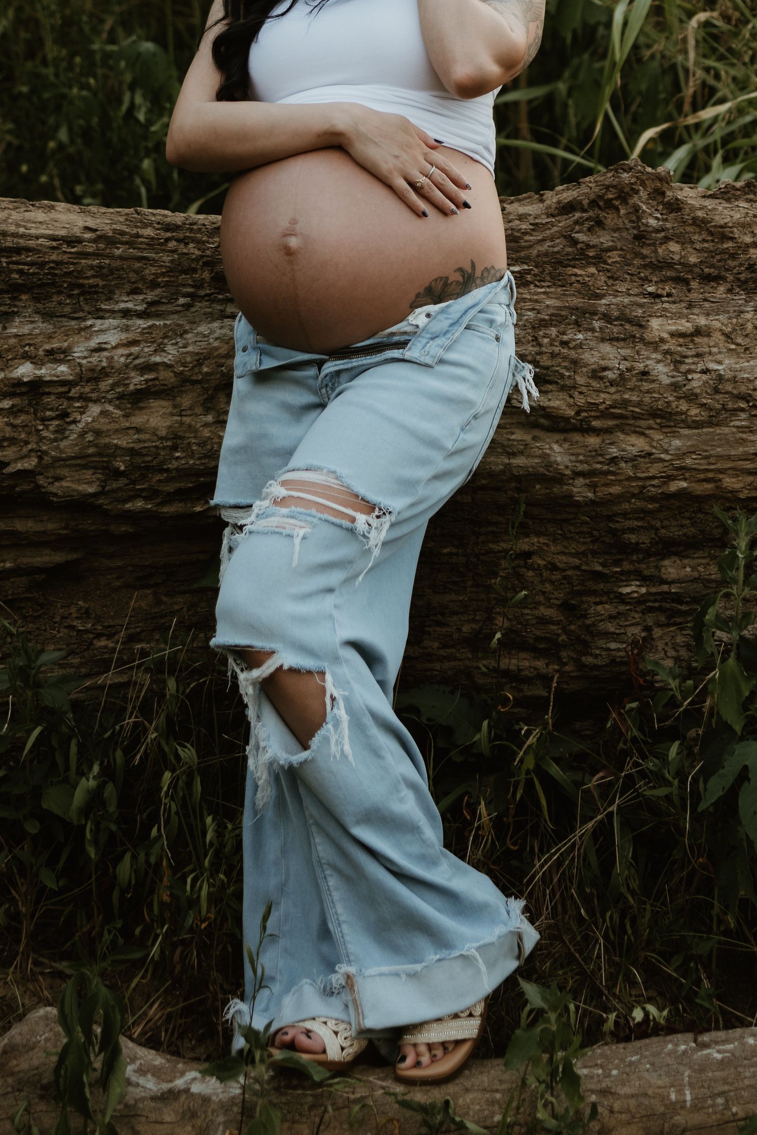 A pregnant belly is shown against distressed light blue jeans while resting on a fallen log in nature.