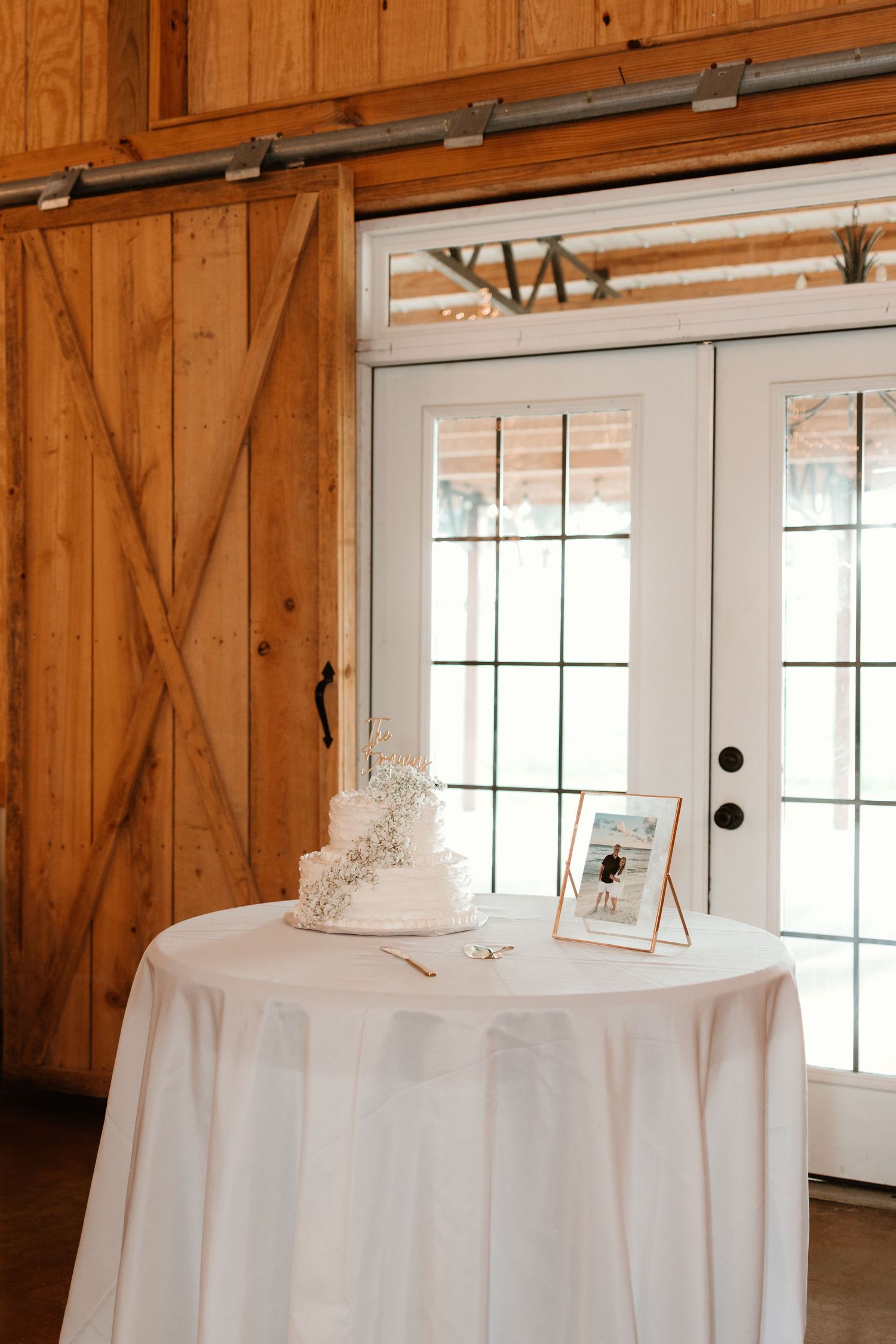 Event venue interior with wooden barn walls and white-draped tables arranged along French doors with natural lighting.
