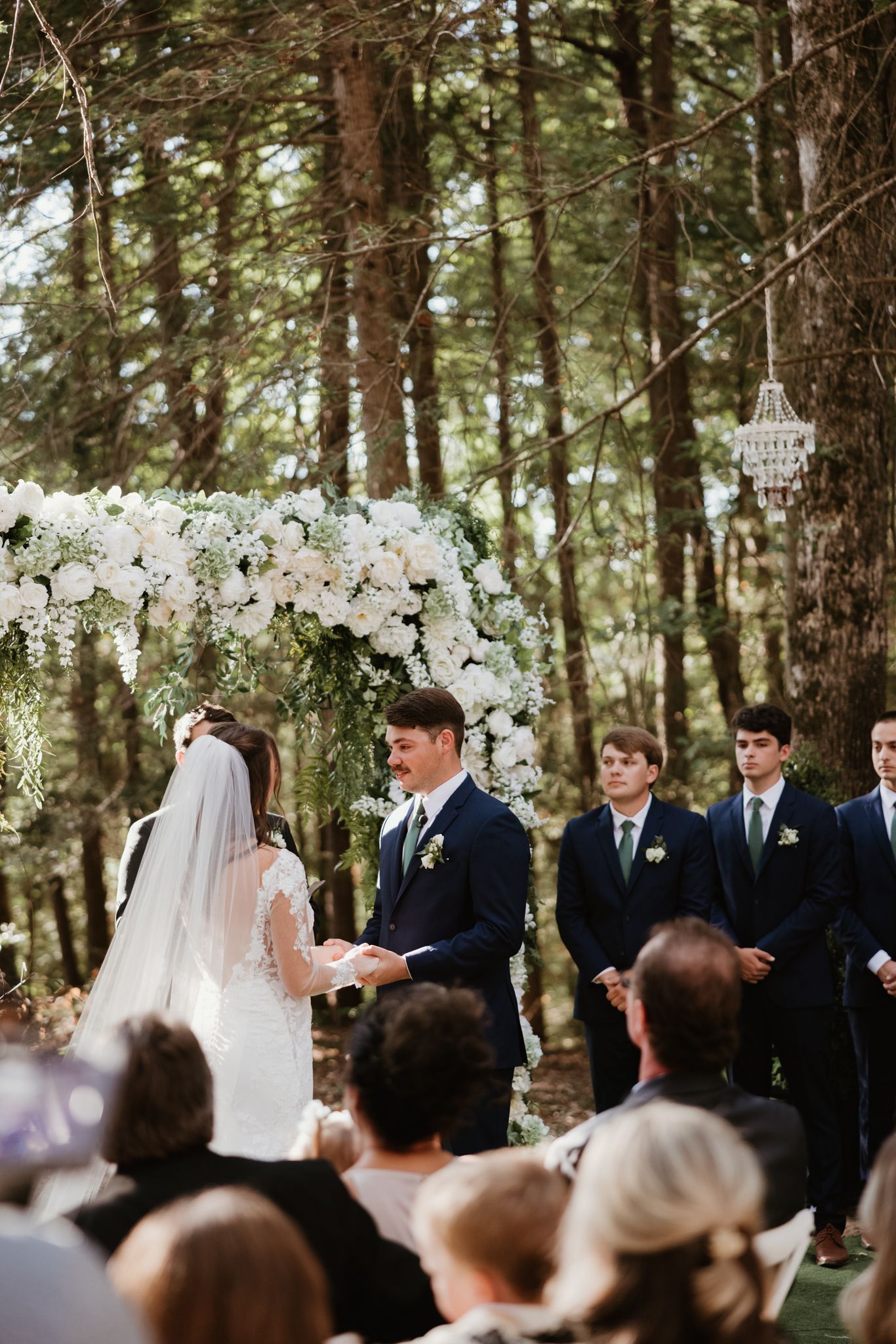 An outdoor forest wedding ceremony with a floral arch and gathered guests.