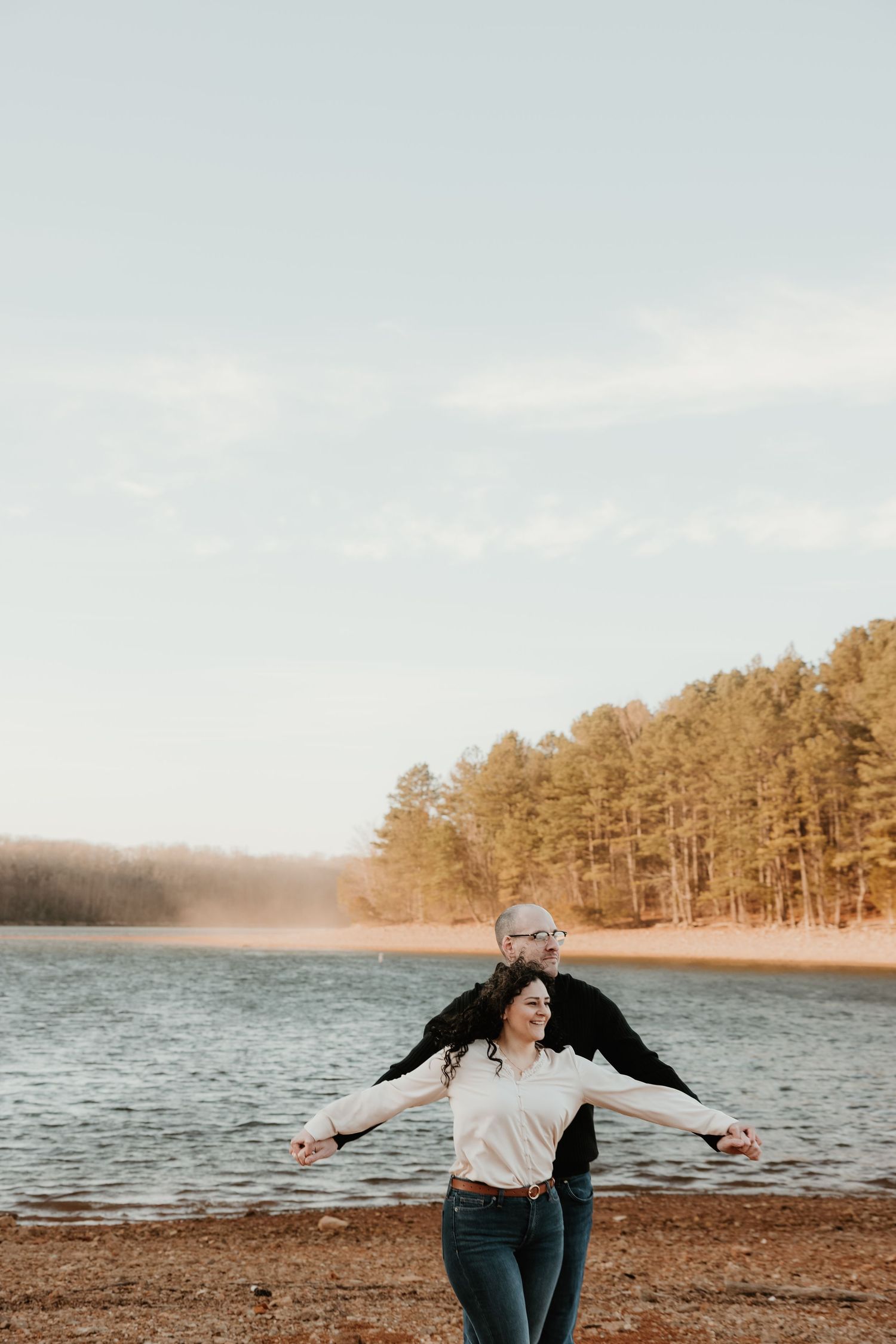 Person with black wings posing playfully by a lake with pine trees in background.