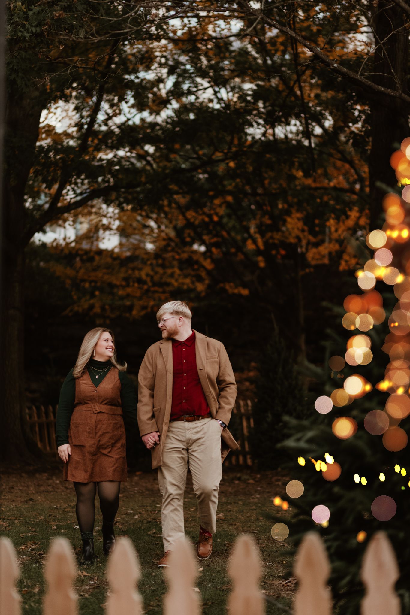 A couple walks hand in hand at dusk beside a white picket fence with bokeh lights glowing in the background.