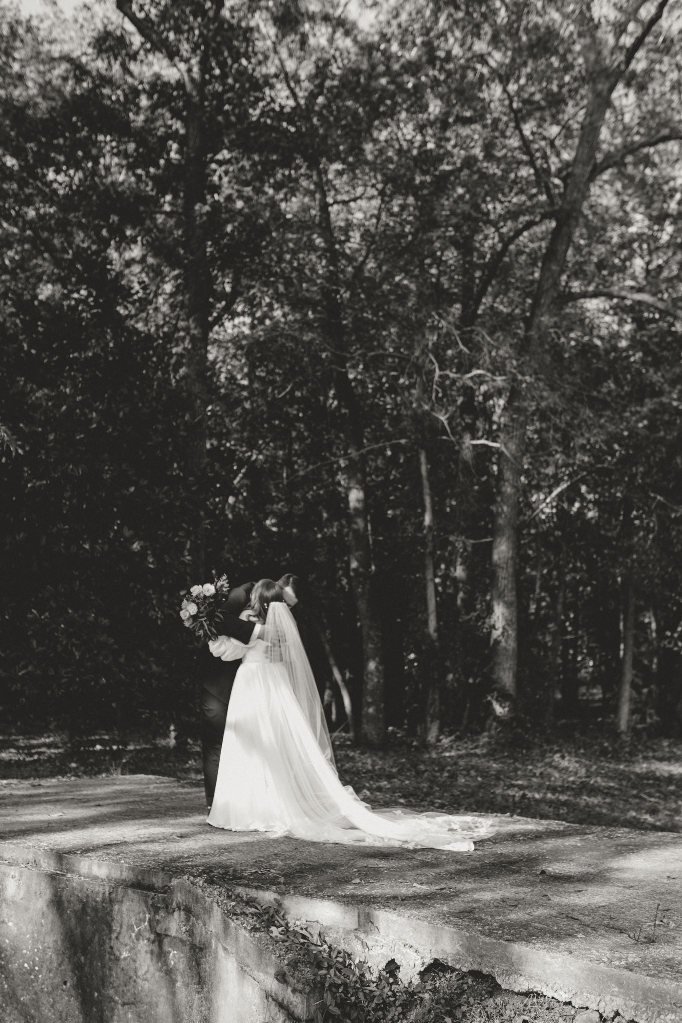 Black and white wedding photo of a bride in long flowing dress and veil standing by stone wall in wooded setting.