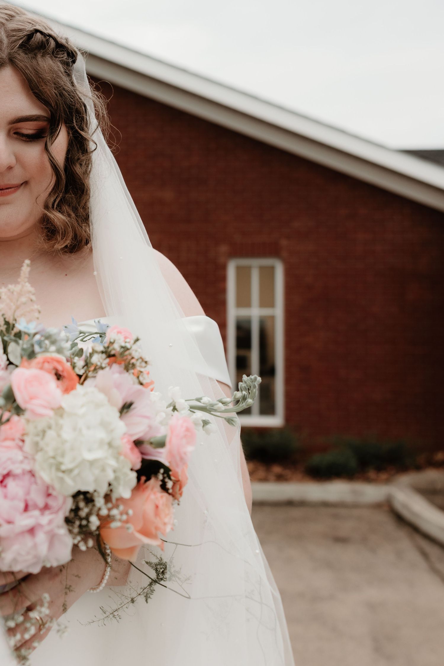 A bride in a white wedding dress holds a beautiful bouquet of pink and white flowers against a red brick building.