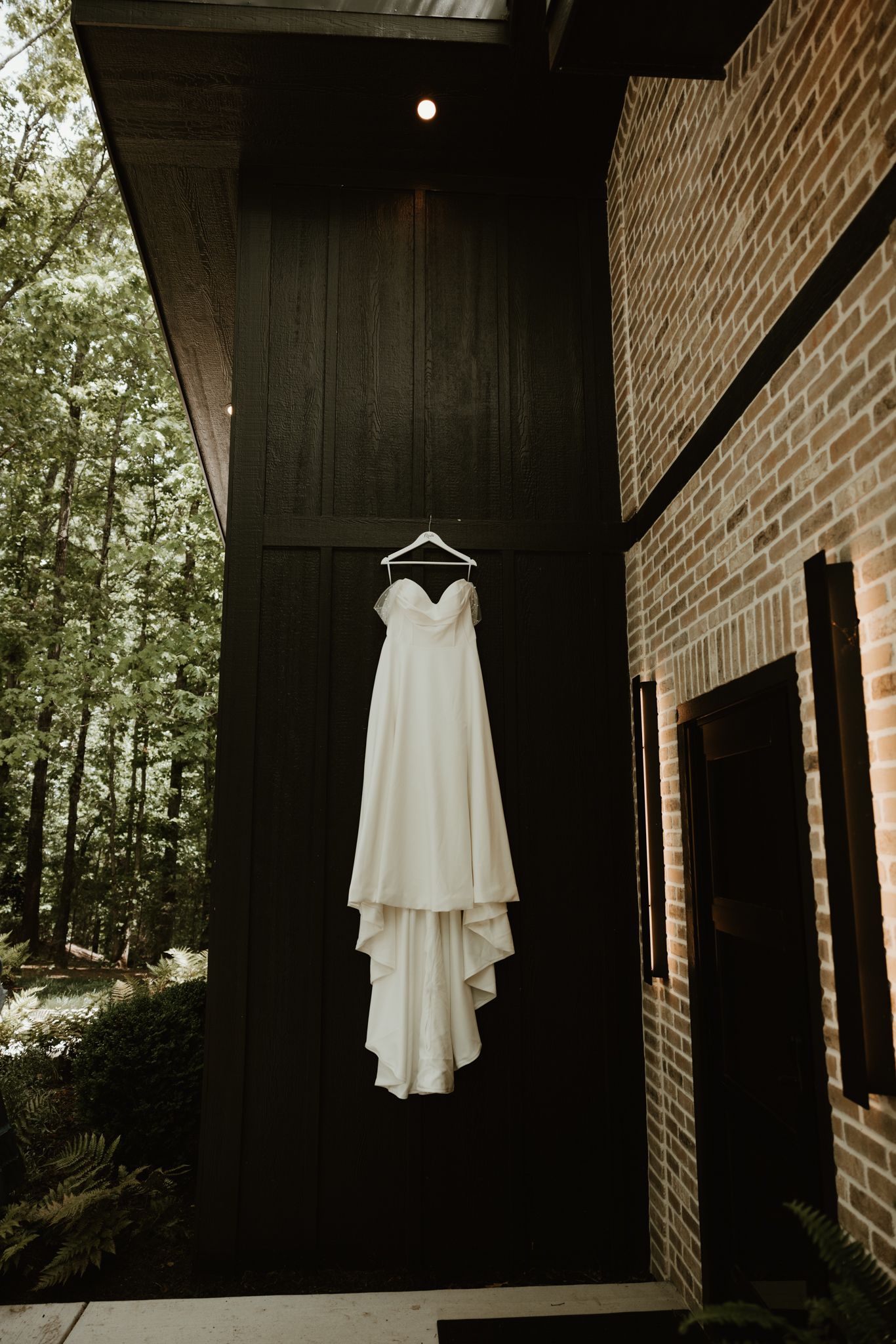 An elegant white wedding dress suspended against a dark wall of a rustic brick building.